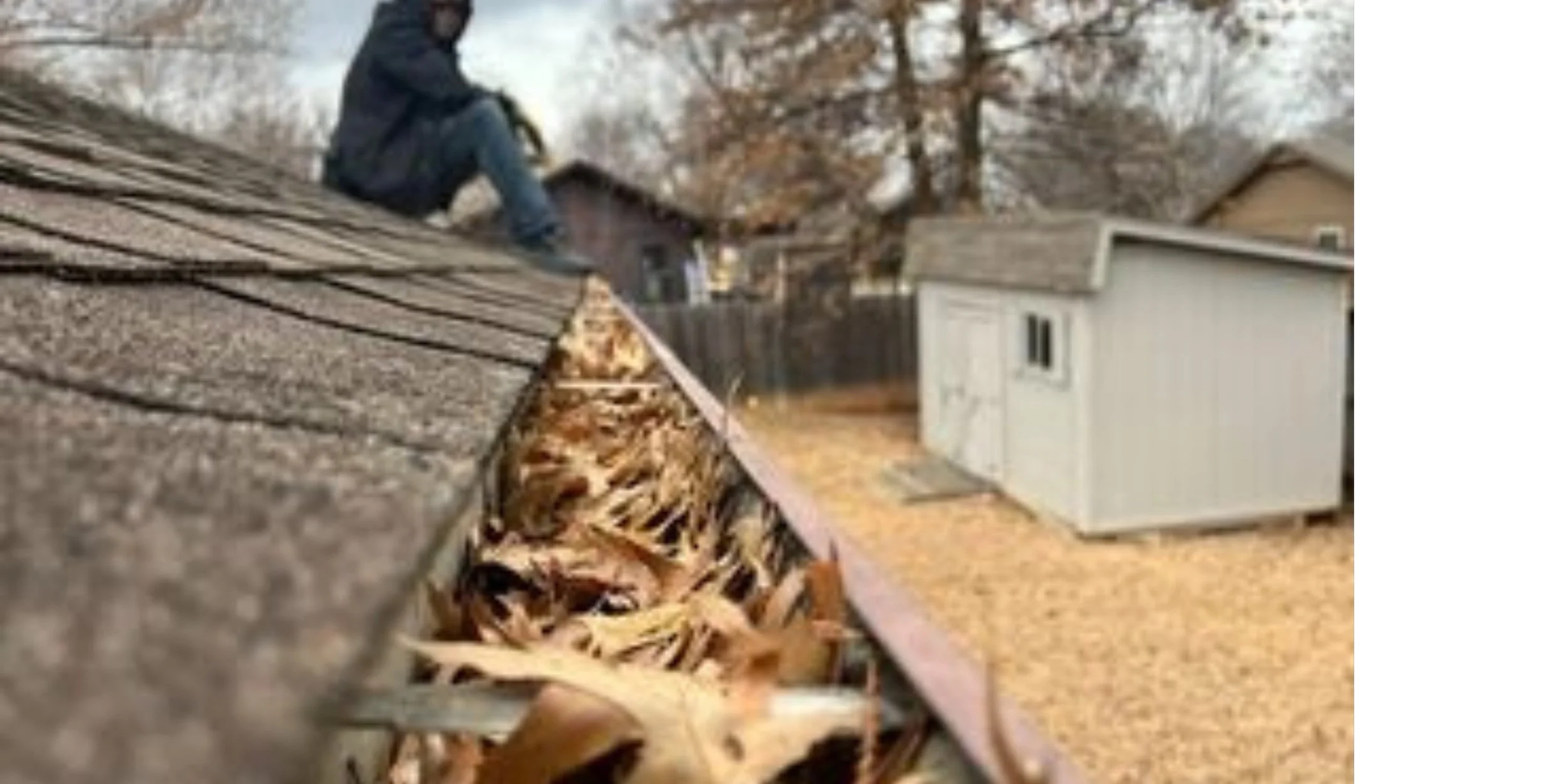 A person working on a roof with shingles, surrounded by trees and houses, during autumn.