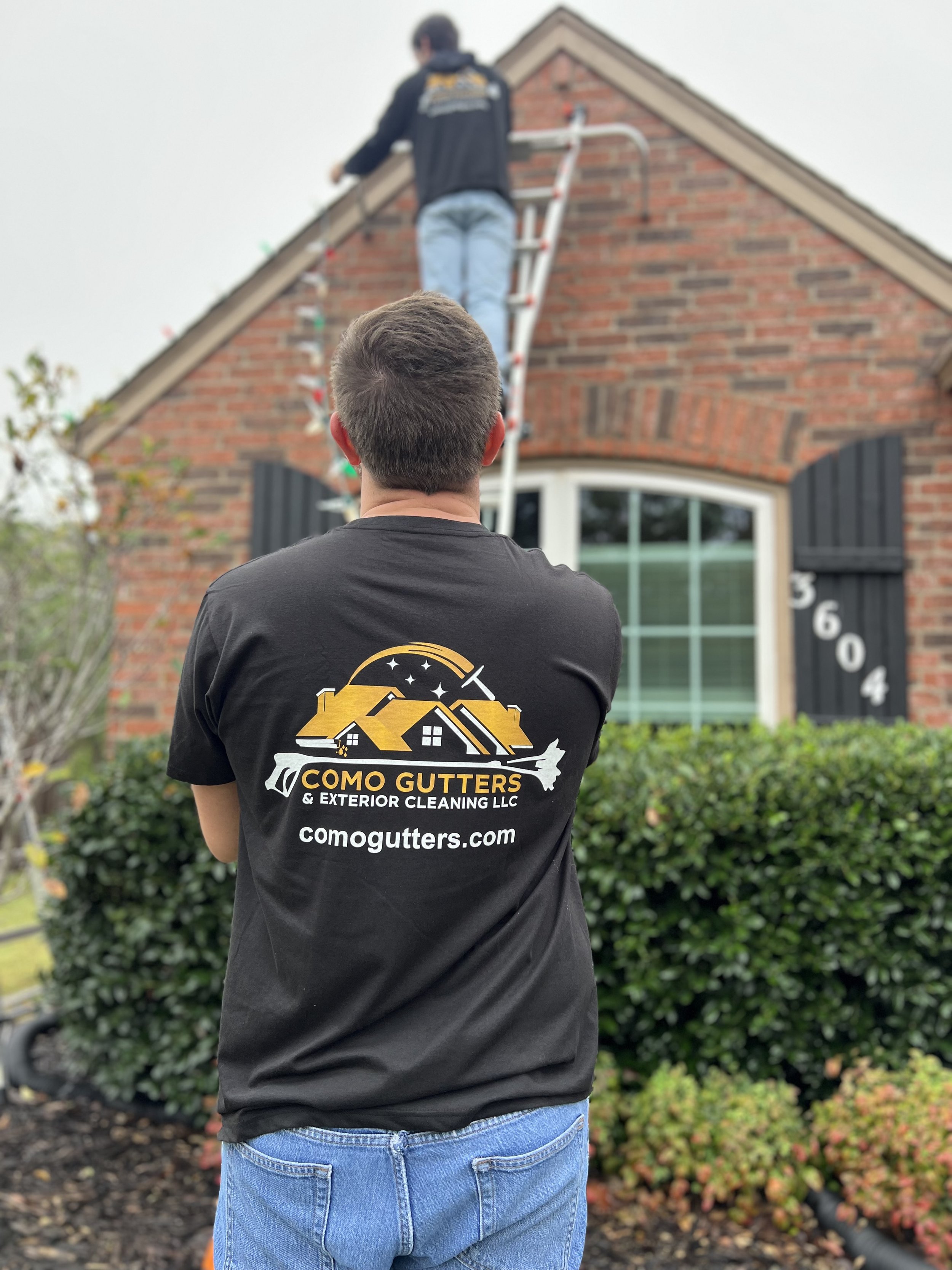 A person with gray hair, wearing a black T-shirt and blue jeans, stands outdoors with their back to the camera, facing a house with a brick facade. Another person is on a ladder working on the house's roof.
