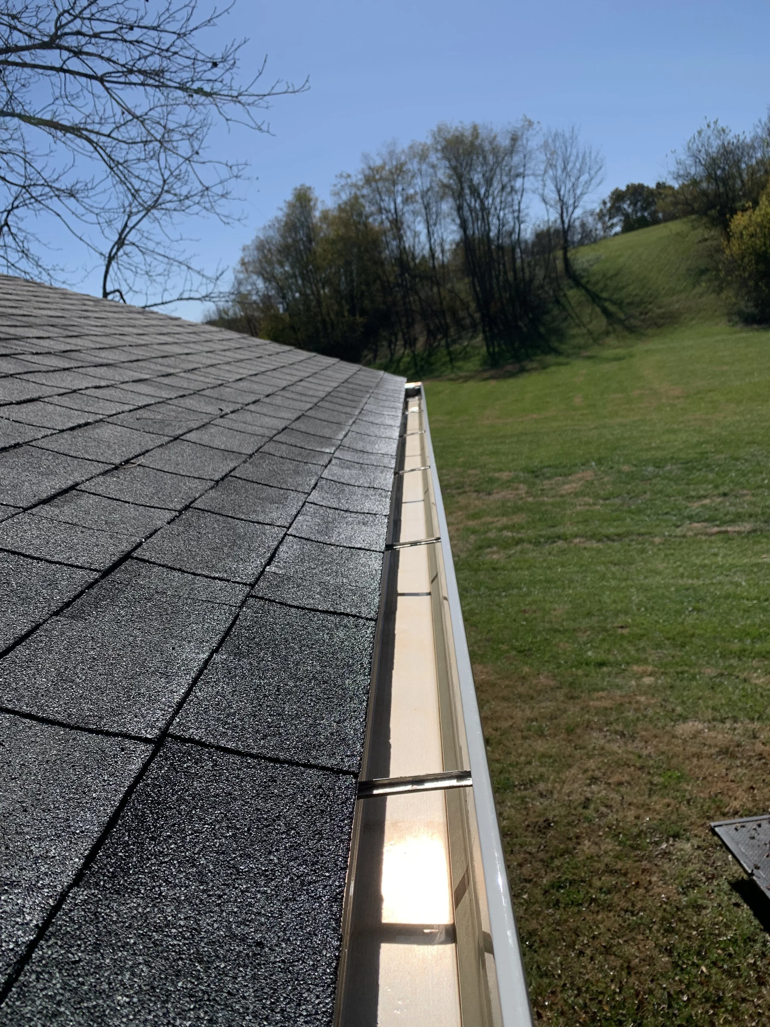 Close-up of a residential house's shingled roof with black asphalt shingles, showing the eaves and gutter system, with a grassy hillside, trees, and blue sky in the background.