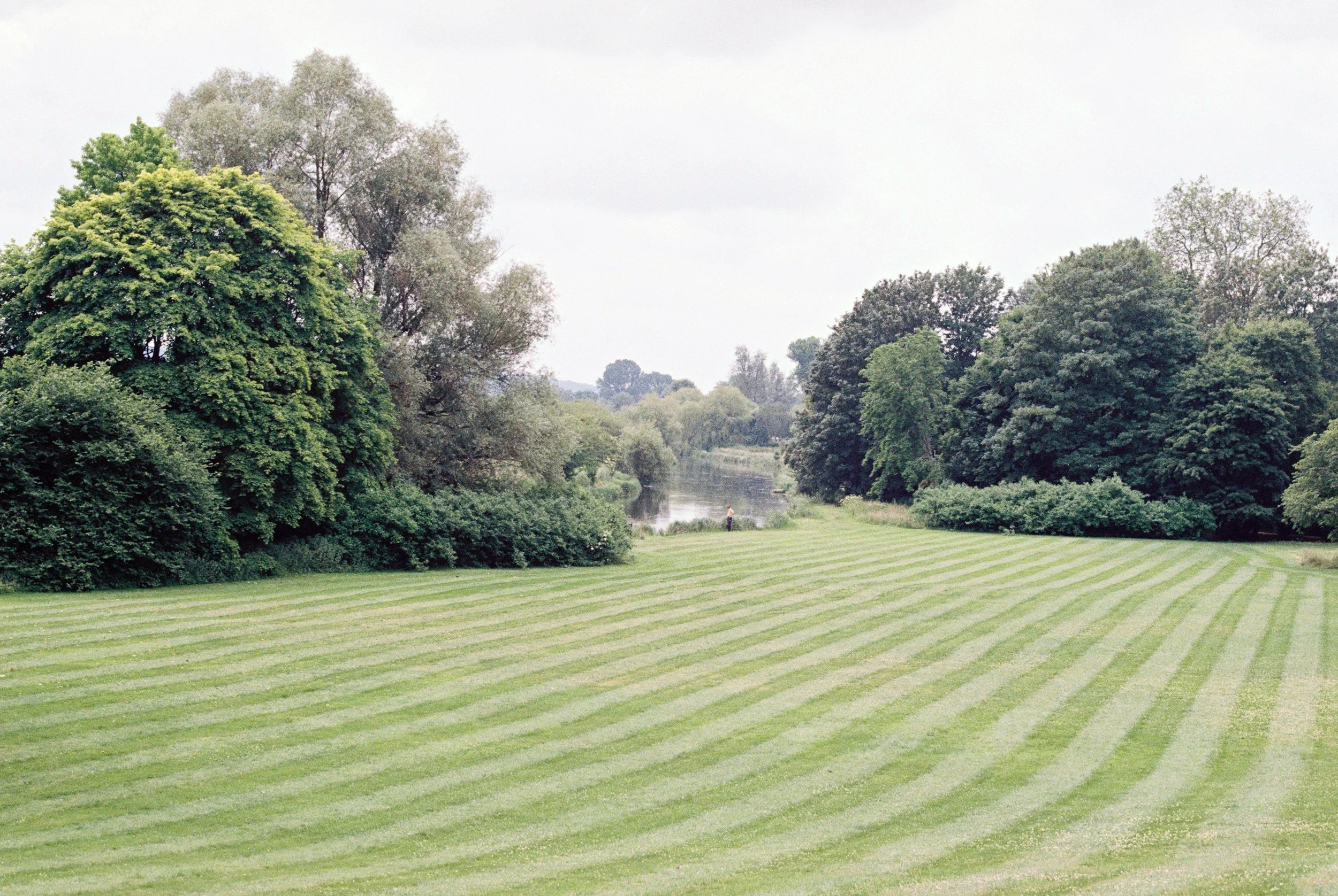 A vast grassy field with lawn stripes, bordered by dense green trees and a river in the distance.