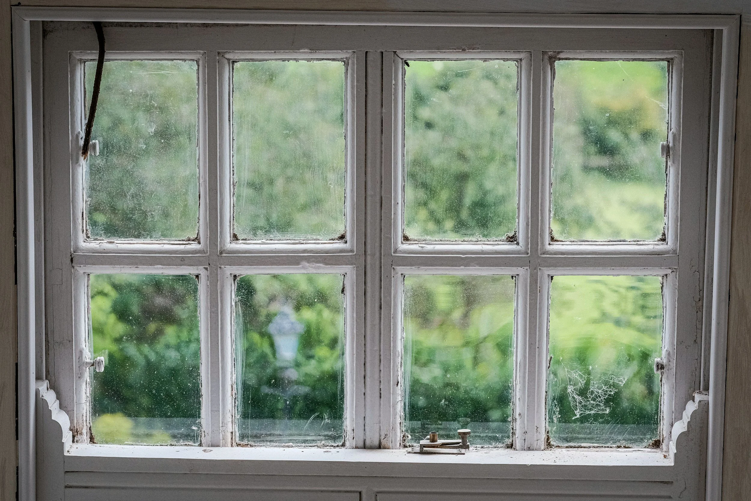 Old, dusty window with multiple panes and cobwebs, looking out onto a green, blurred outdoor scene.