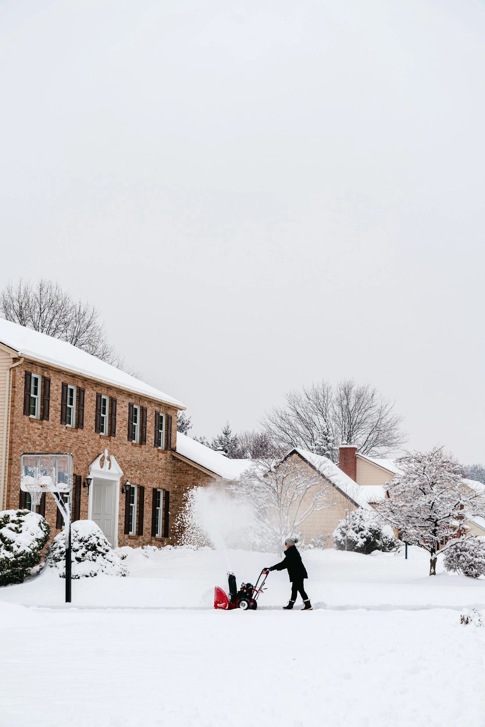 A man clearing snow with a snowblower in a front yard of a brick house with four windows and a white door during winter, with snow-covered trees and other houses in the background.