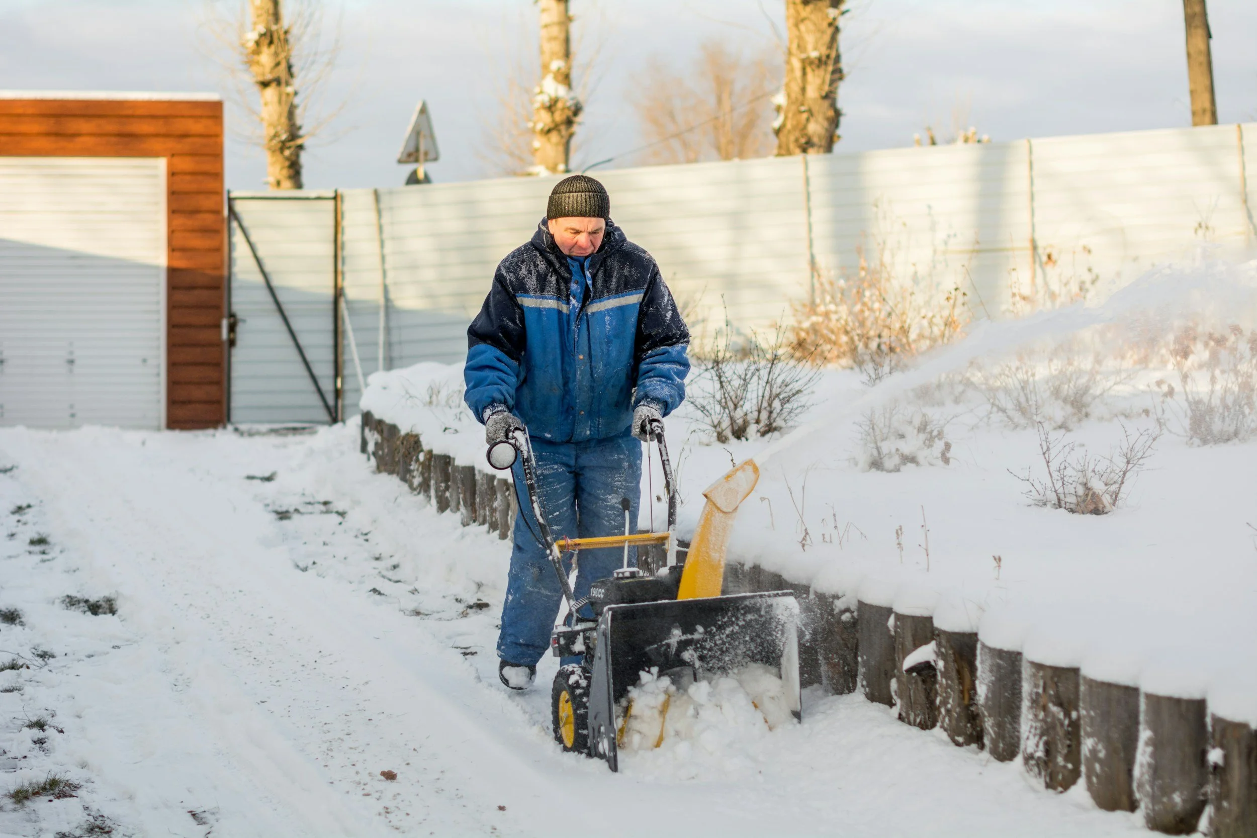 Man in winter clothing using snow blower to clear snow from a sidewalk in a backyard with snow-covered ground, wooden fence, and trees in the background.