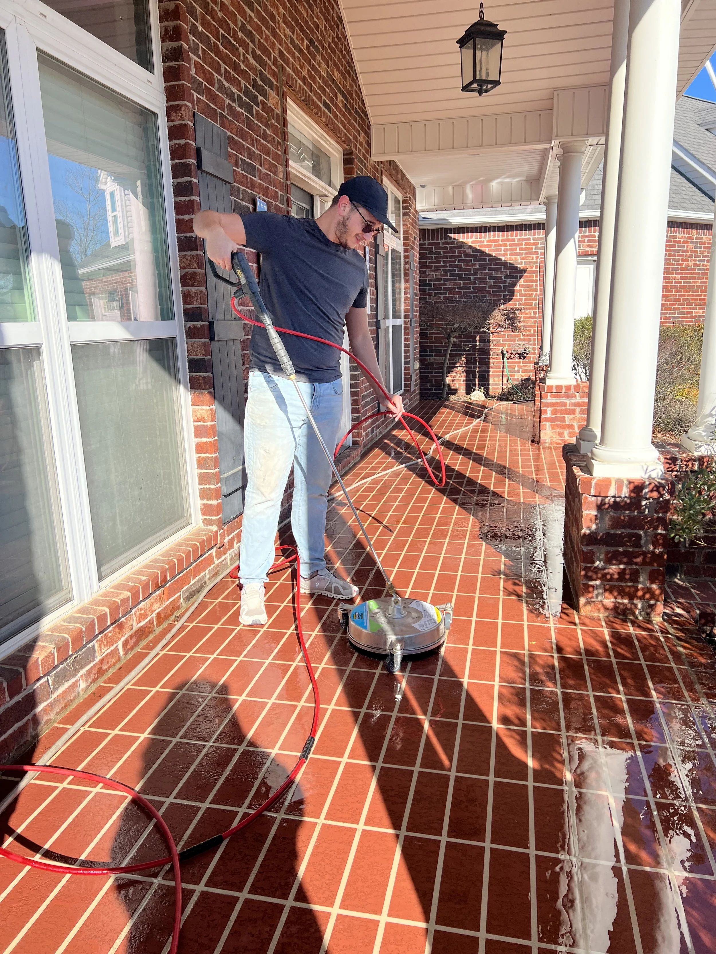 A man in dark clothing, sunglasses, and a cap is pressure washing a brick porch with a red hose attached to a surface cleaner. The porch is attached to a brick house with white columns and large windows, and the man is standing on the cleaned, wet surface.