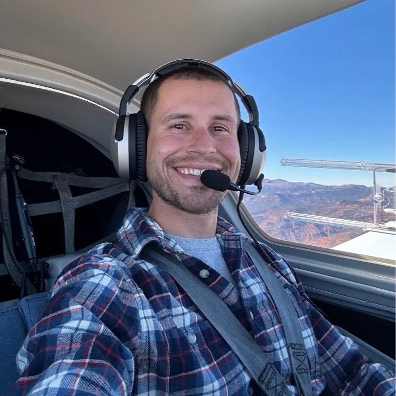 Man flying a small aircraft, smiling, wearing a headset and plaid shirt, inside cockpit with mountain landscape visible through window