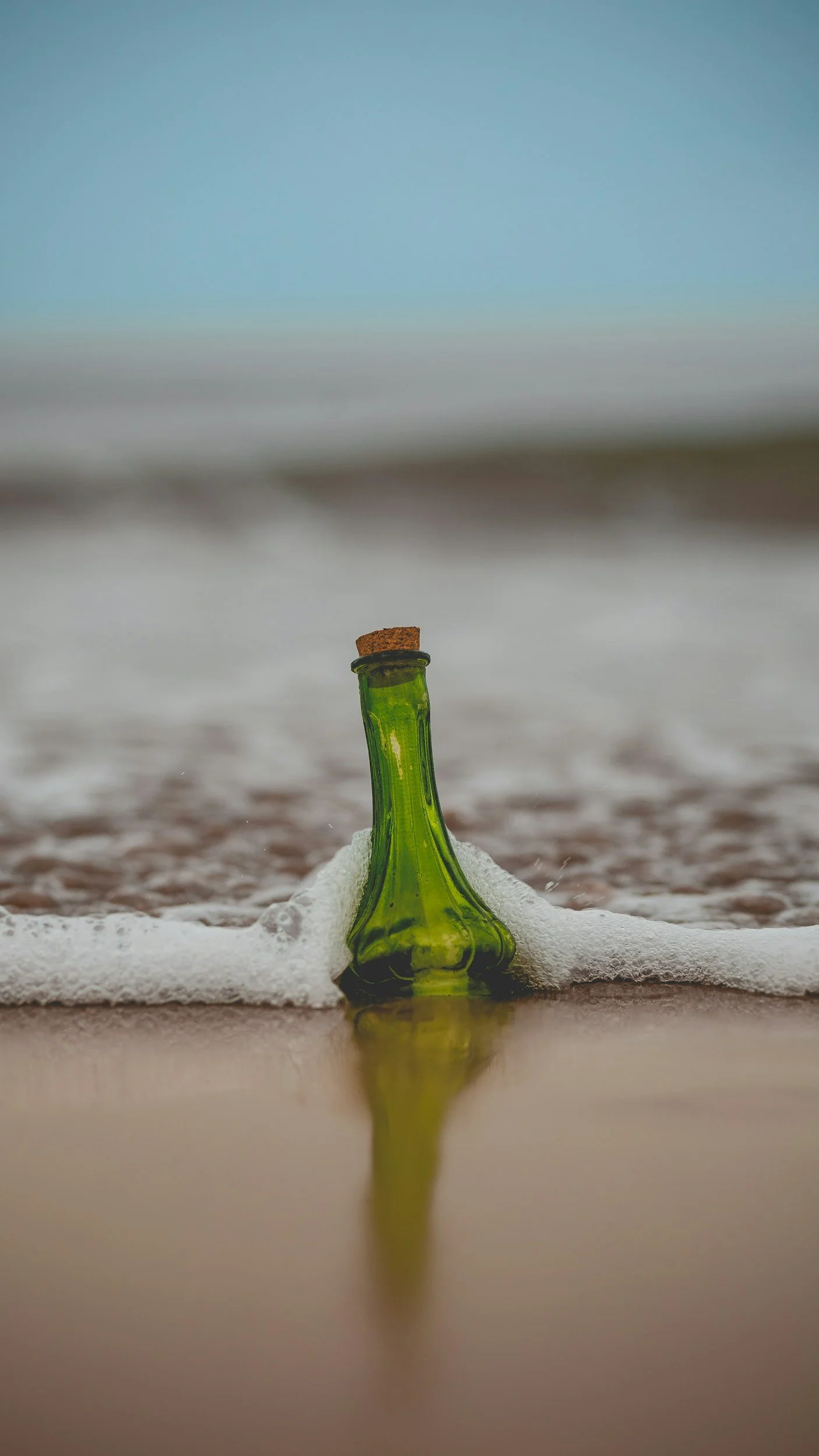 A green glass bottle with a cork in the neck, partially buried in the sand on a beach, with its reflection visible on the wet sand, surrounded by foamy waves, under a blue sky.