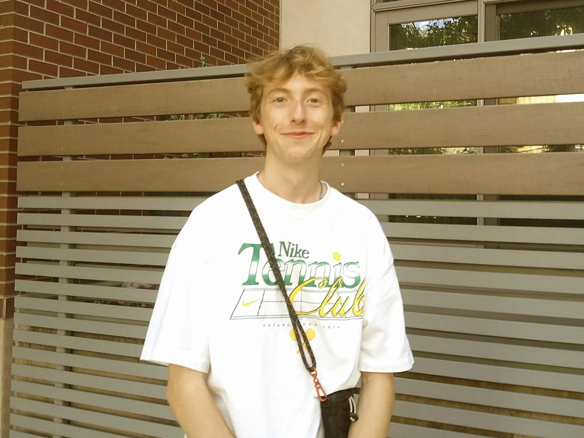 A young man with curly blonde hair stands outside in front of a wooden slat fence and a brick building, smiling at the camera. He is wearing a white T-shirt with colorful text and has a black strap across his shoulder.