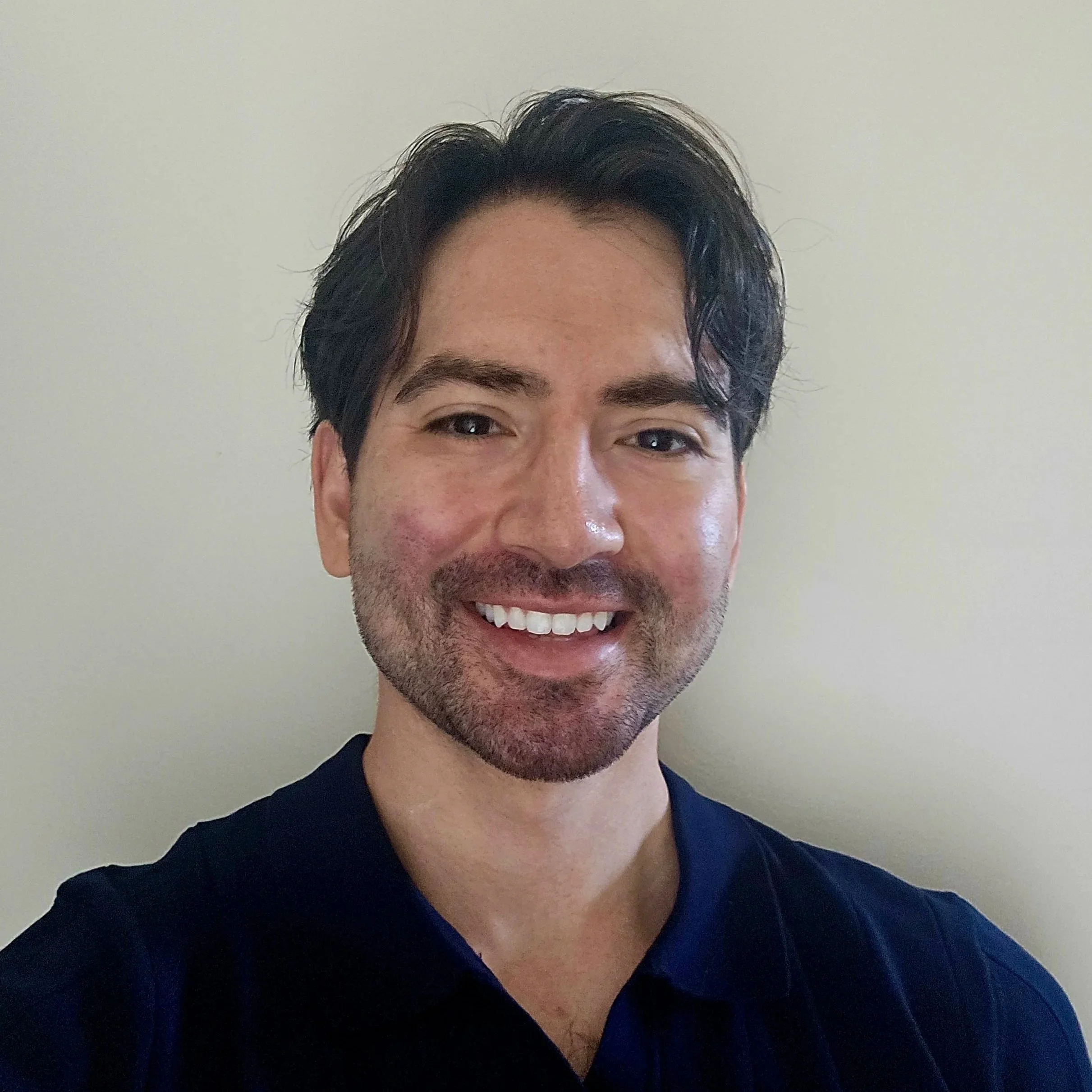 A close-up of a smiling man with dark hair, eyebrows, and facial hair, wearing a navy blue shirt, against a plain light-colored background.