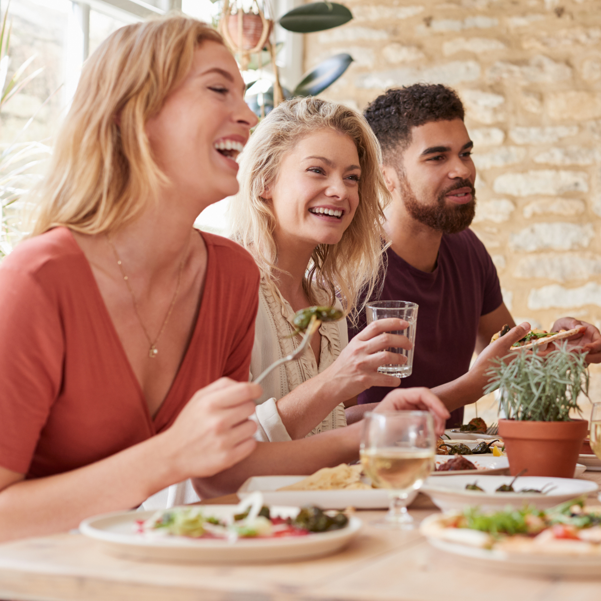 A group of four people smiling and enjoying a meal together at a dining table with various plates of food and drinks, in a bright room with a brick wall background.
