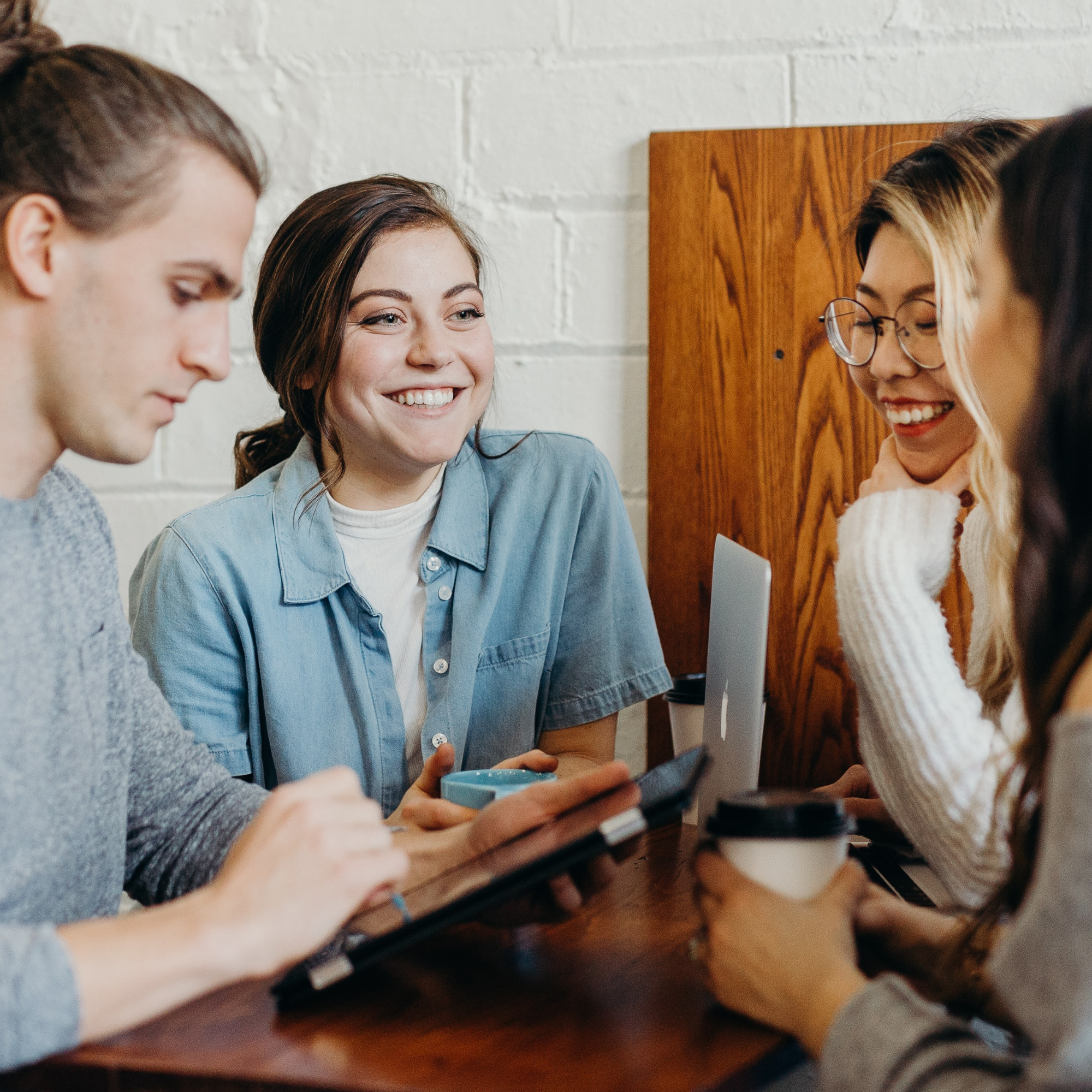 Four young women are gathered around a wooden table, smiling and engaging with a laptop and a smartphone.