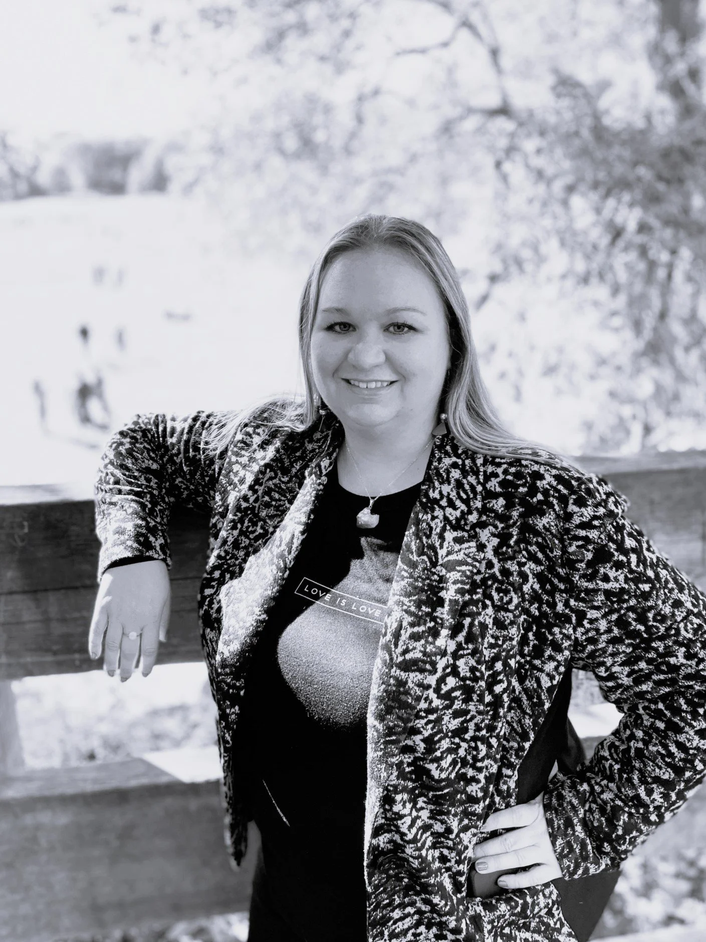 A black and white portrait of an artist, TBARNES CREATES, in a patterned blazer, leaning with one arm on a fence, and a city park in the background.