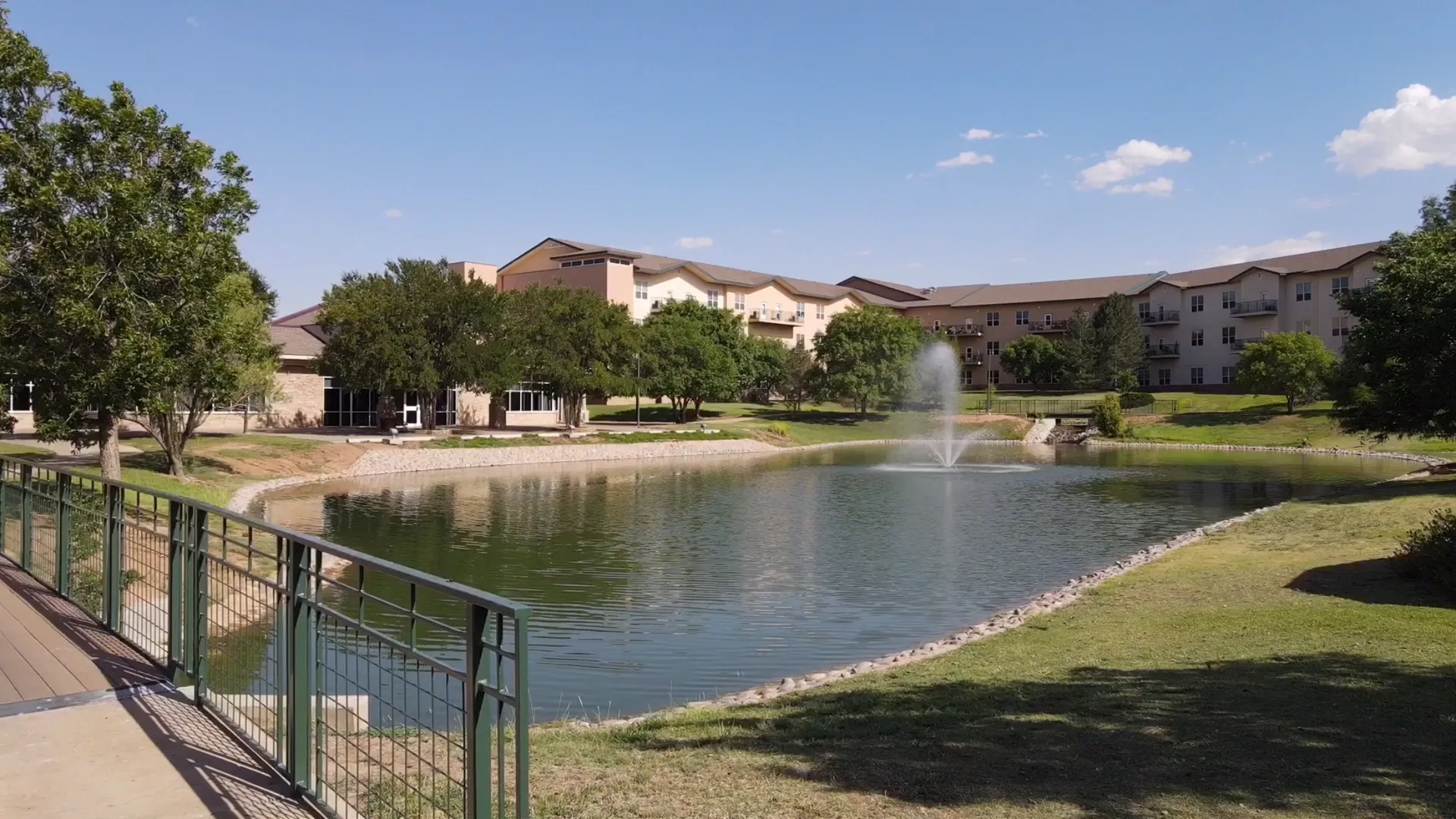 A pond with a fountain in the middle, surrounded by trees and a grassy area, with multiple apartment buildings in the background. A walkway with a green railing runs along the pond's edge.