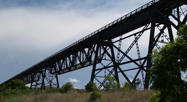 Steel railroad bridge crossing over a grassy area with trees, under a partly cloudy sky.