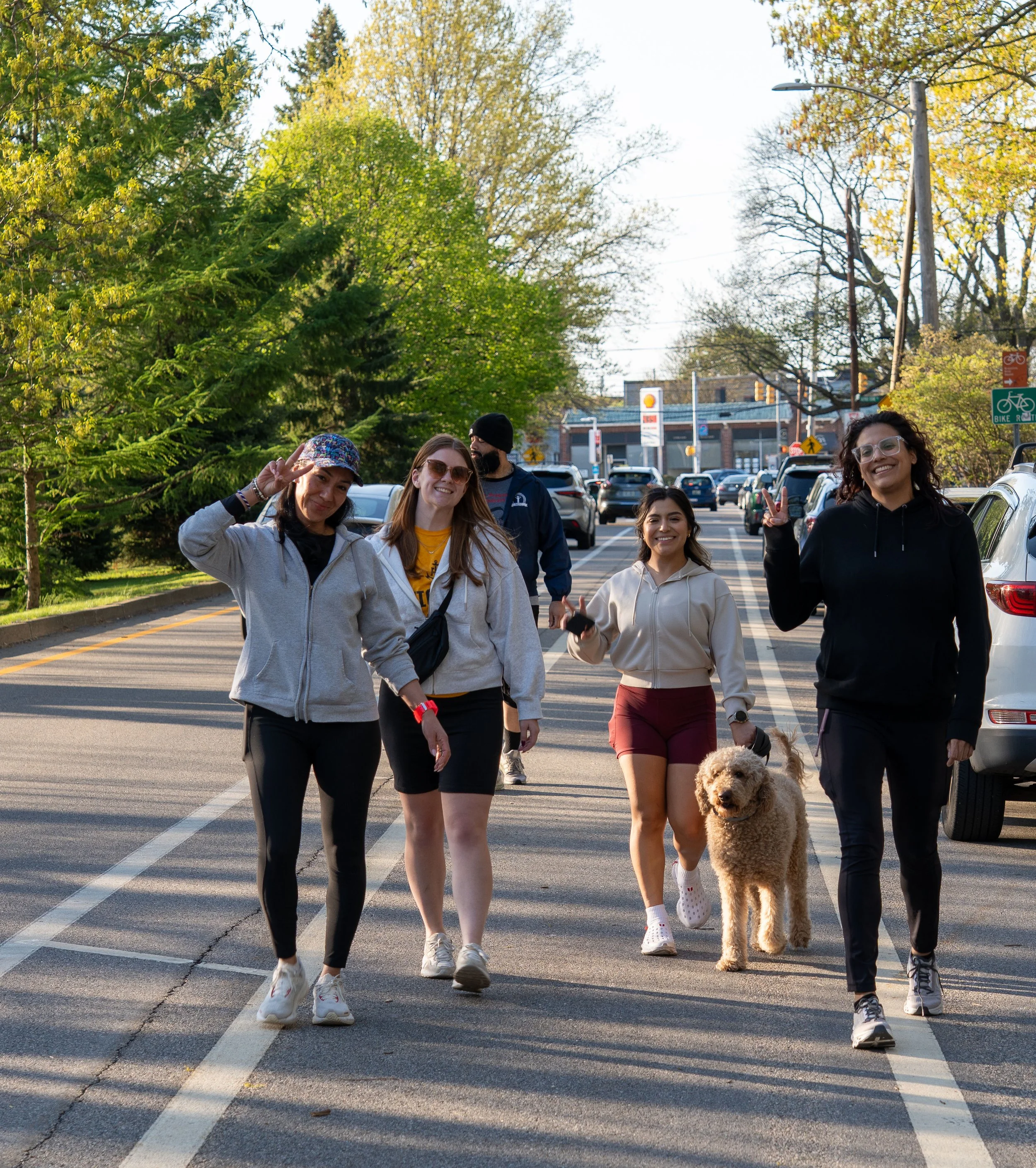 A group of five people and a dog walking across a parking lot on a sunny day with trees and store buildings in the background.