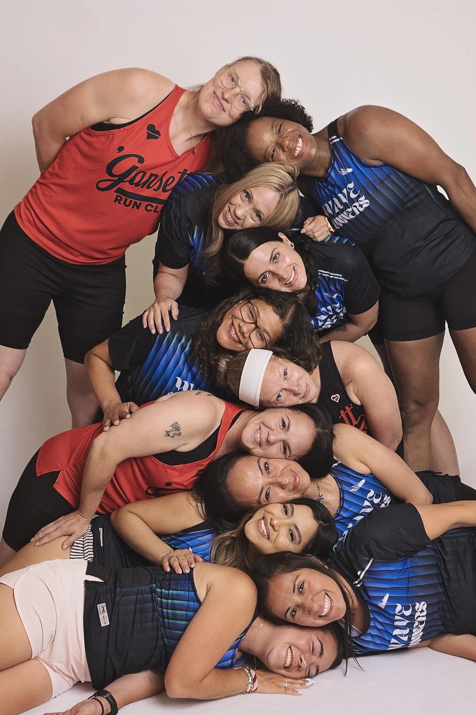 Group of women in sportswear lying on the floor, smiling and forming a human pyramid against a plain light-colored background.