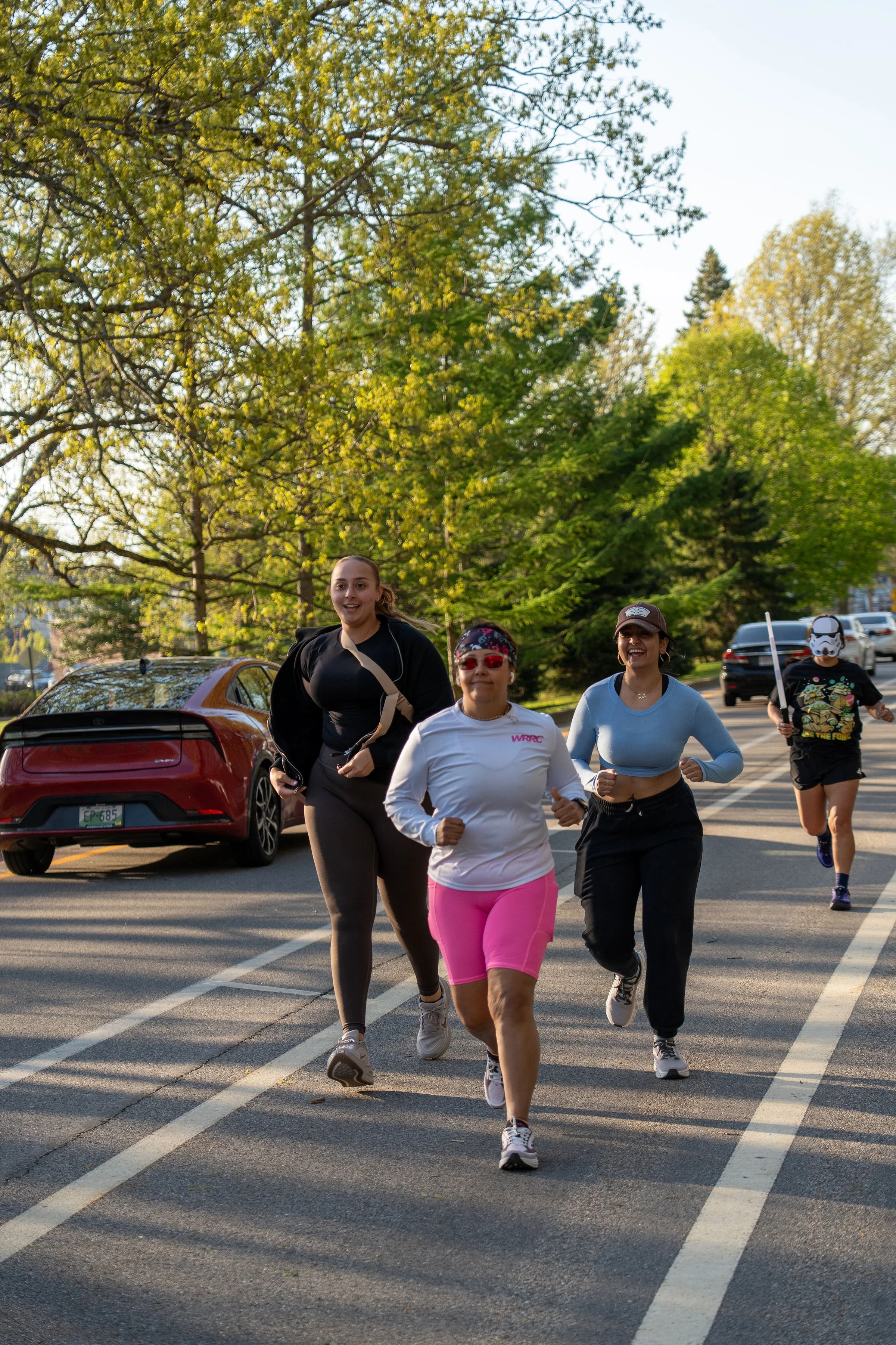 A group of women jogging outdoors in a parking lot with trees and parked cars in the background.