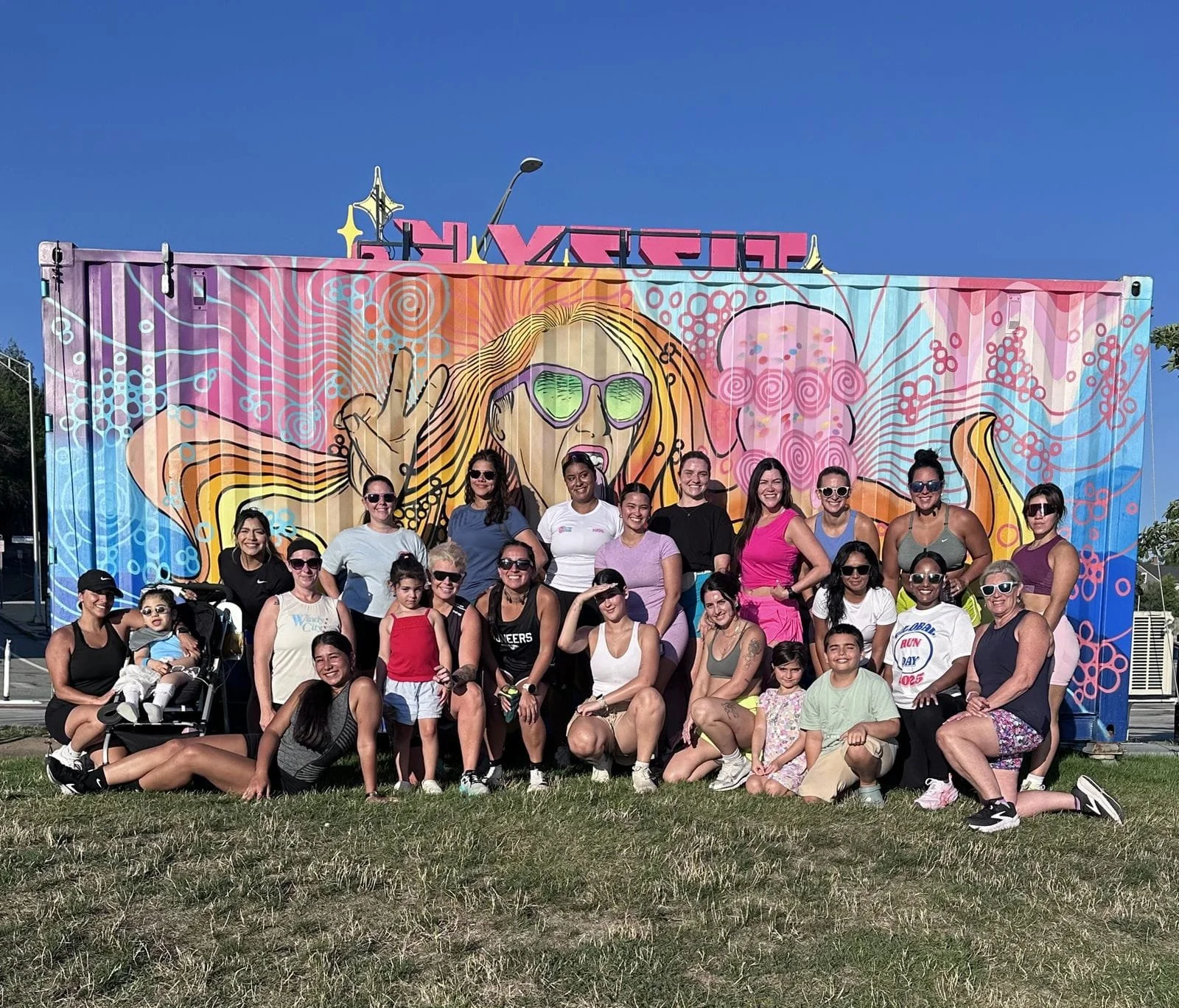 A group of women and children posing in front of a colorful mural of a woman with sunglasses, long hair, and making a peace sign with her hand.