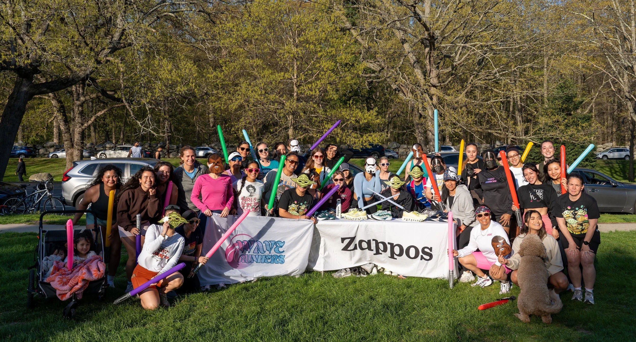 A large group of women gathered outdoors for a Star Wars-themed event, holding toy lightsabers and wearing character masks, standing behind a Zappos banner with trees and parked cars in the background.