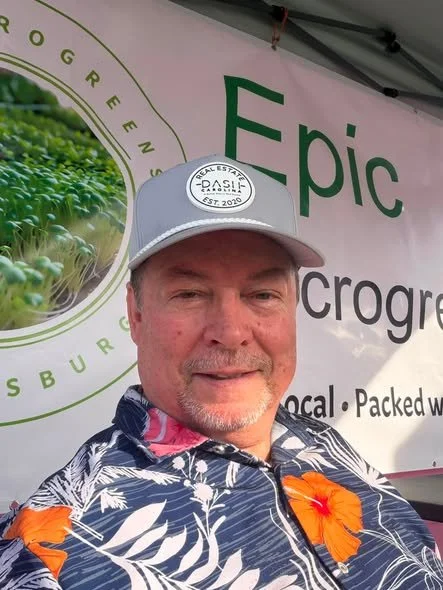 Man wearing a floral shirt and a gray cap with a white circular patch, standing in front of a pink booth with green text that says 'Epic'; part of a green logo with a farm scene is visible on the left.
