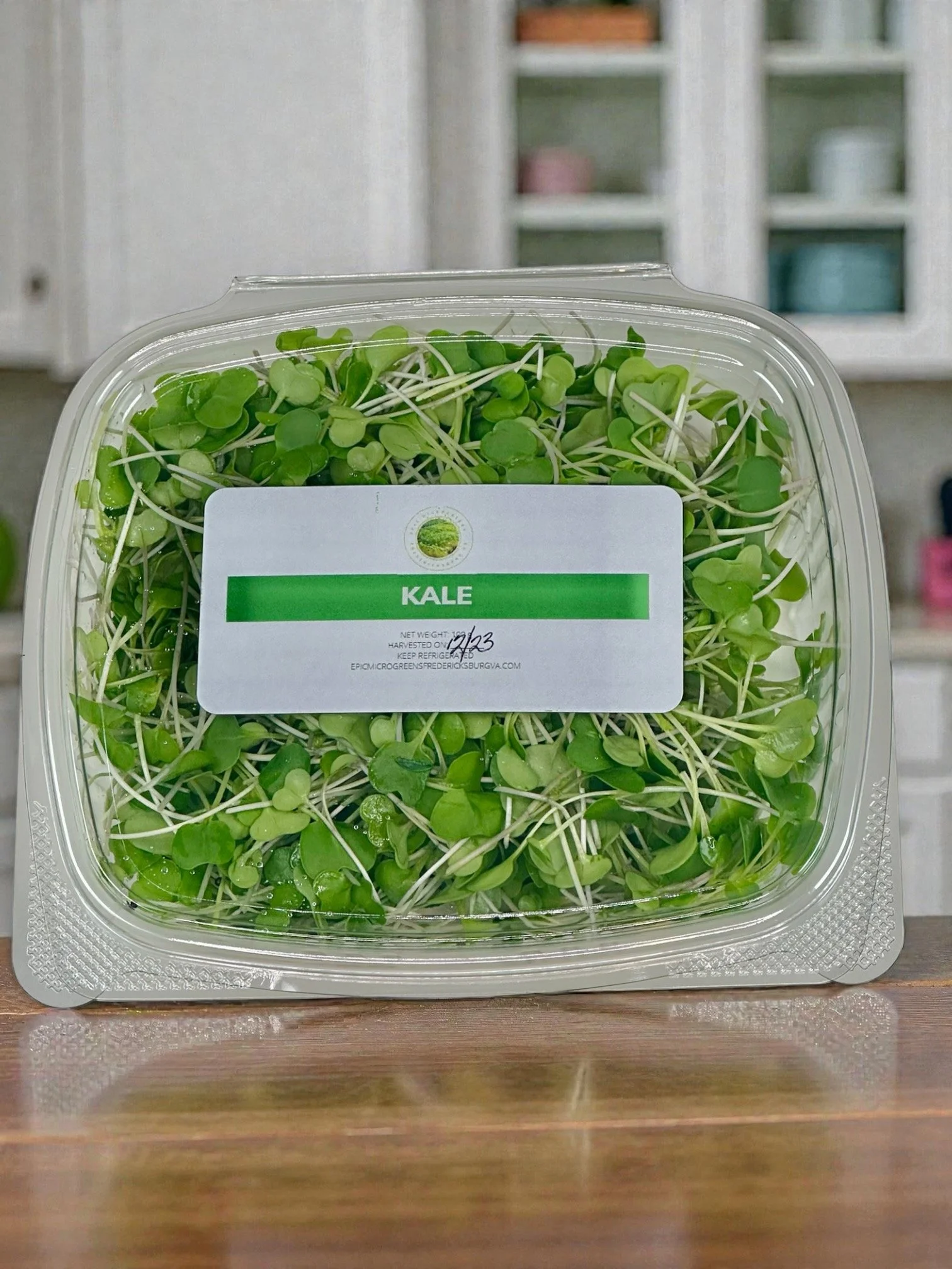 A clear plastic container of microgreens labeled 'KALE' on a wooden surface.