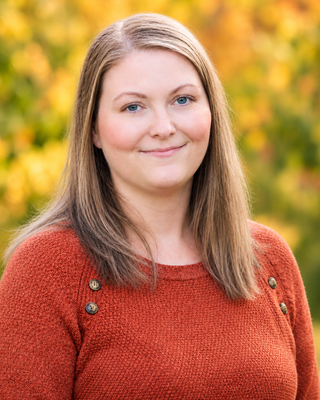 A young woman with long blonde hair wearing a red sweater outdoors with yellow autumn foliage in the background.