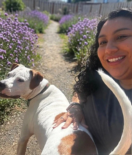 A woman with dark curly hair and her white and tan dog both facing the camera with a path with purple wildflowers behind them.