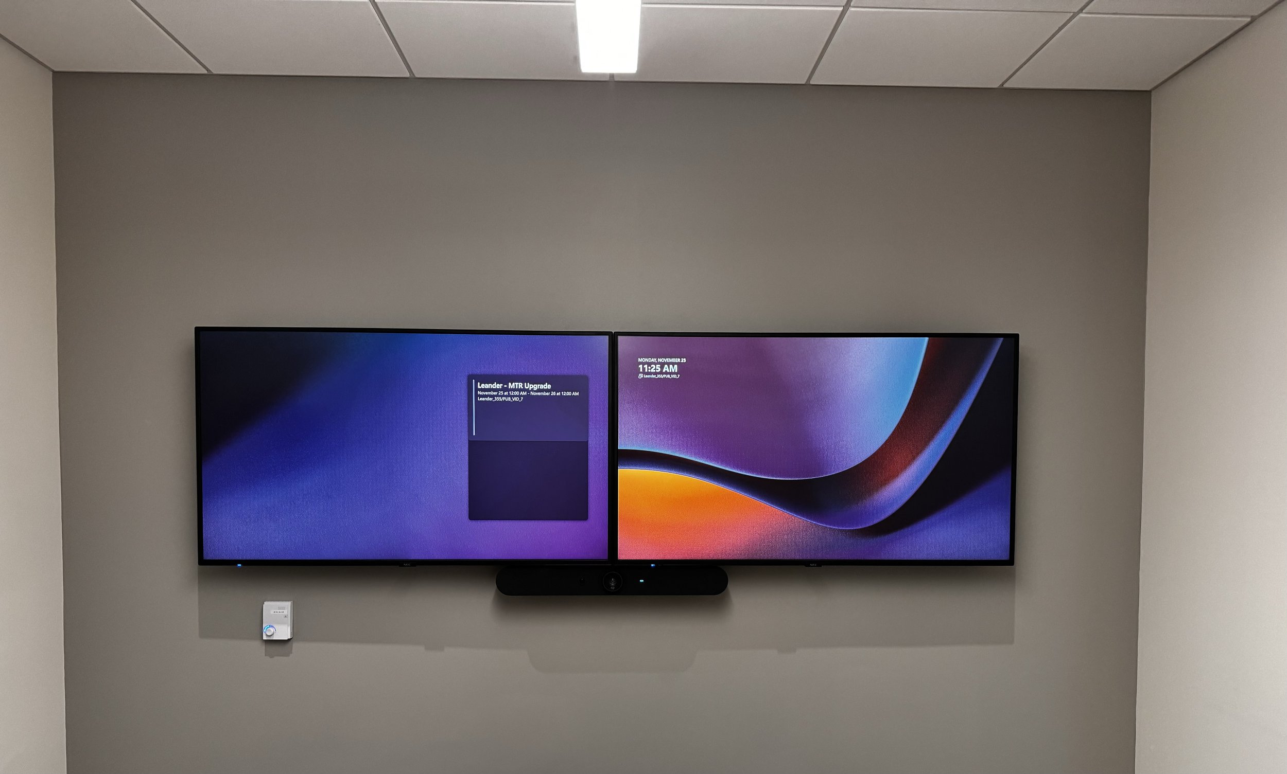 A wall-mounted flat-screen monitor displays a colorful digital background with text indicating a scheduled upgrade, time, date, and network details, located in a room with a gray wall and a ceiling with rectangular tiles.
