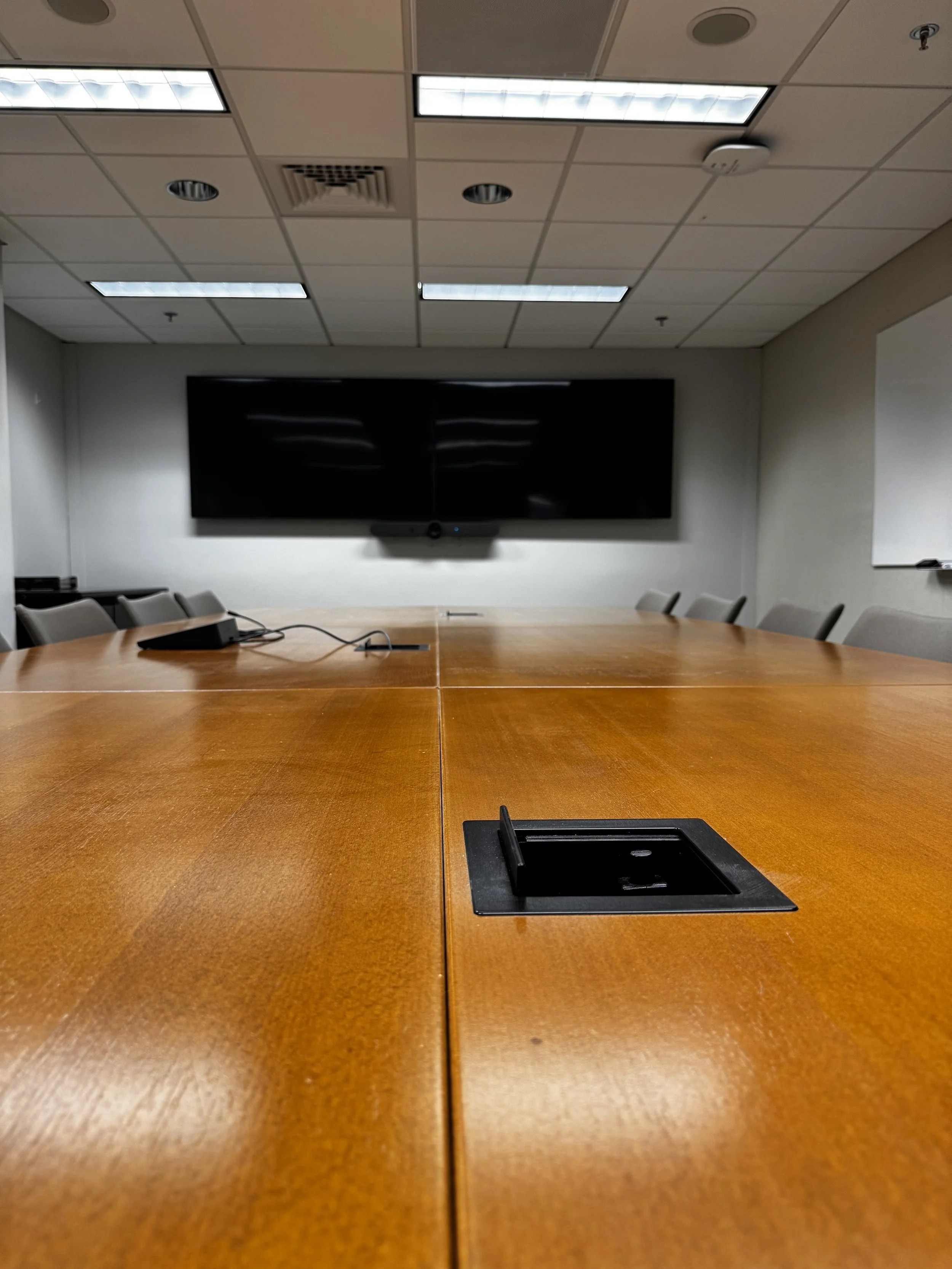 Conference room with wooden table, chairs, a wall-mounted large black screen, a whiteboard, and ceiling lights.