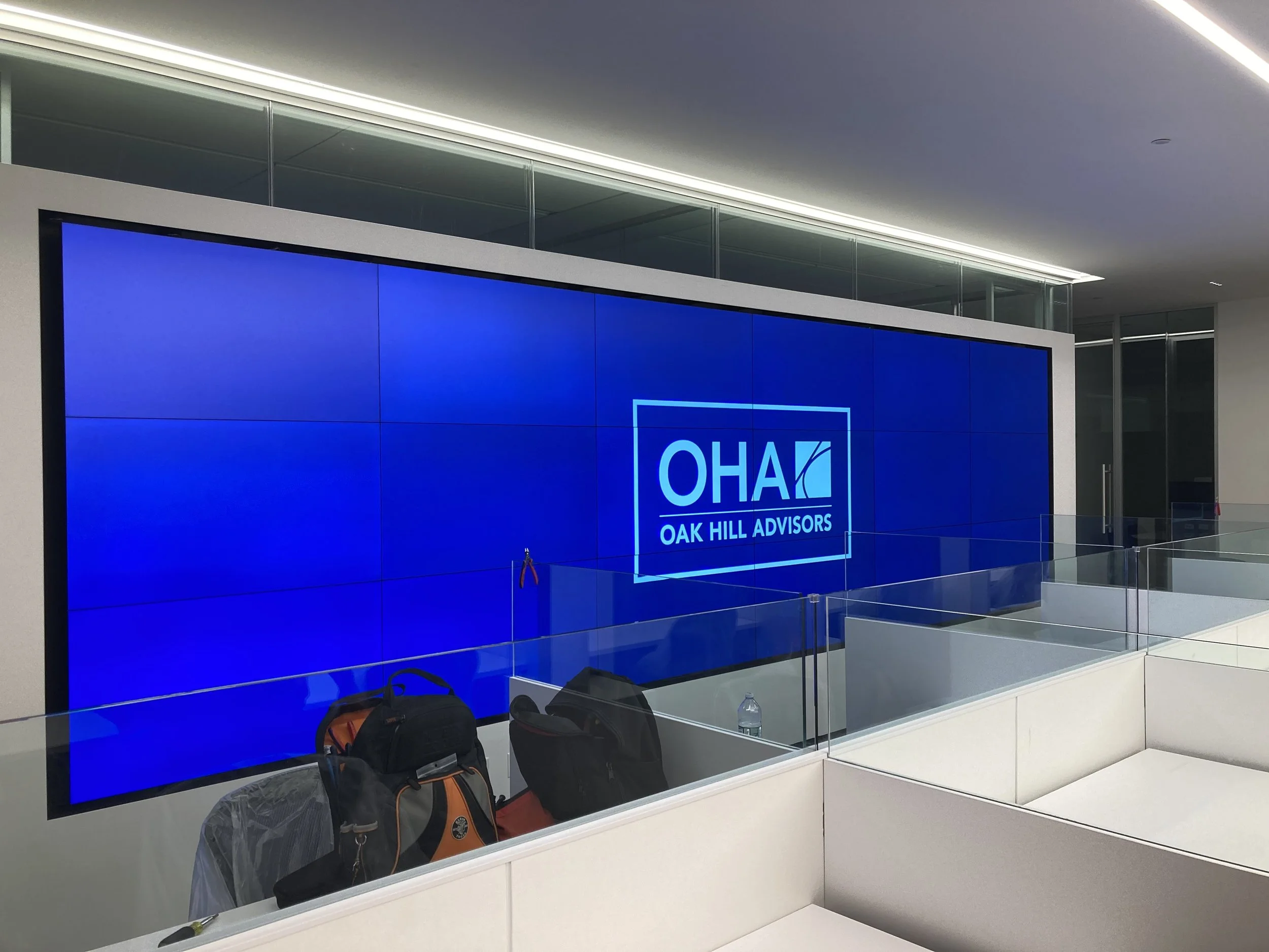 Large digital screen displaying the logo and name of OAK HILL ADVISORS in an modern office lobby with a glass wall overhead. There are multiple backpacks and a water bottle on the ledge in front of the screen.
