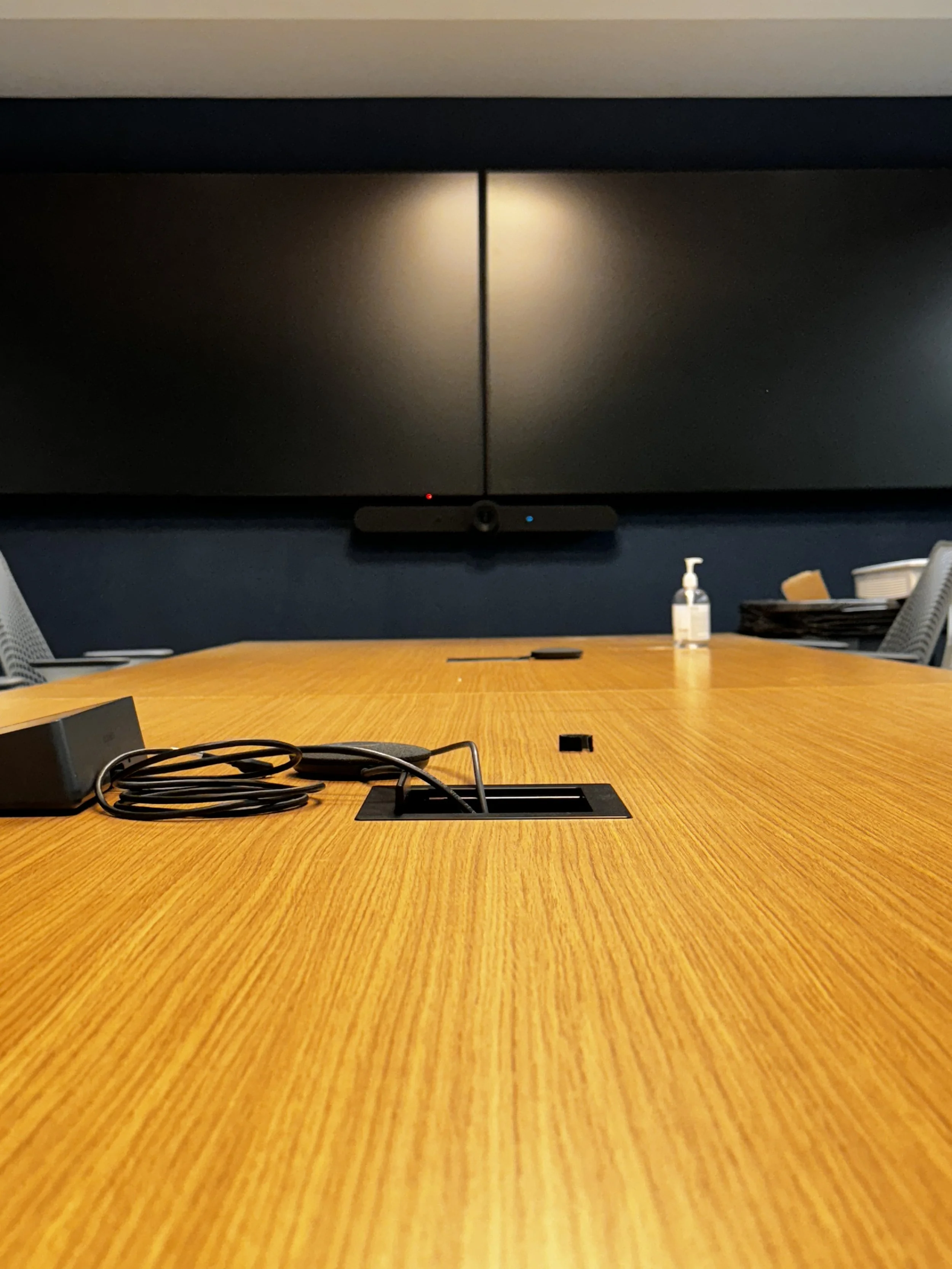 A conference room table with a black conference phone, a bottle of hand sanitizer, and some cables. A large black screen is mounted on the wall behind.