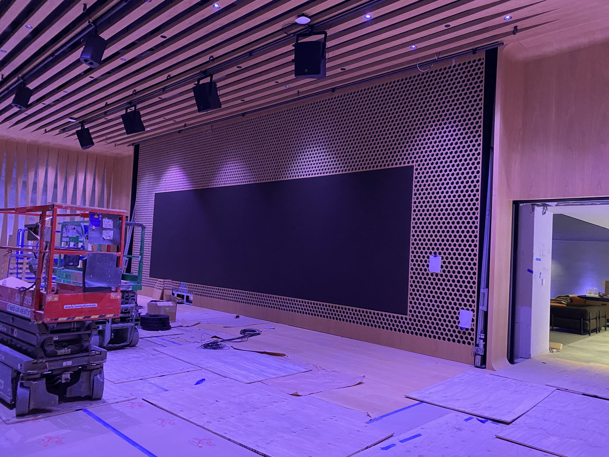 Interior of a room under construction with a large mounted perforated wooden wall panel, ceiling mounted speakers, and construction equipment on the floor.