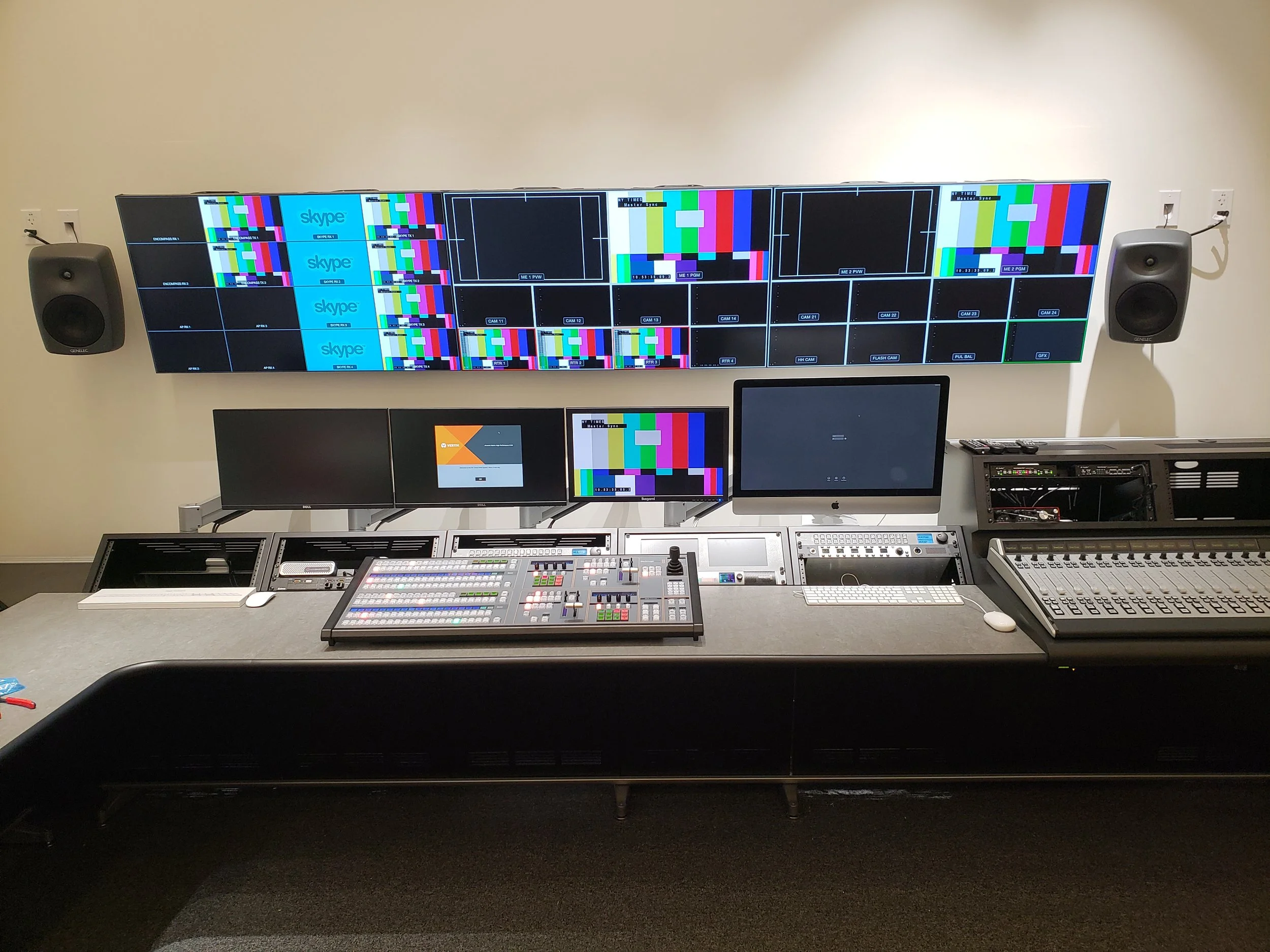 A large video control room with multiple monitors displaying color test patterns and Skype logos, with speakers on either side and various control panels and equipment on the desk.