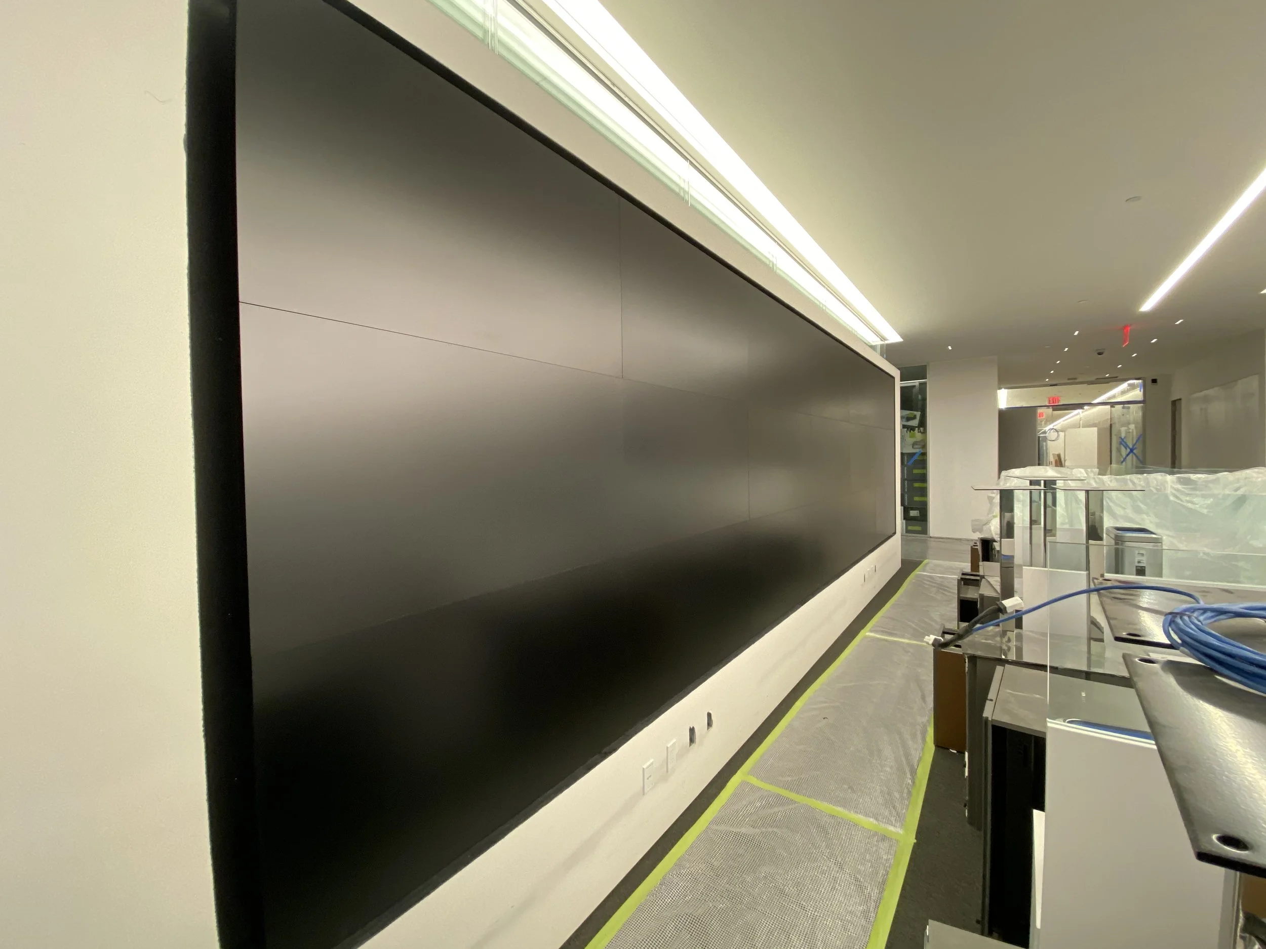 The image shows an indoor construction or setup area with a large black flat panel or screen on the left. To the right, there are construction materials and equipment, including a metal cart, wires, and partially visible plastic covering. The ceiling