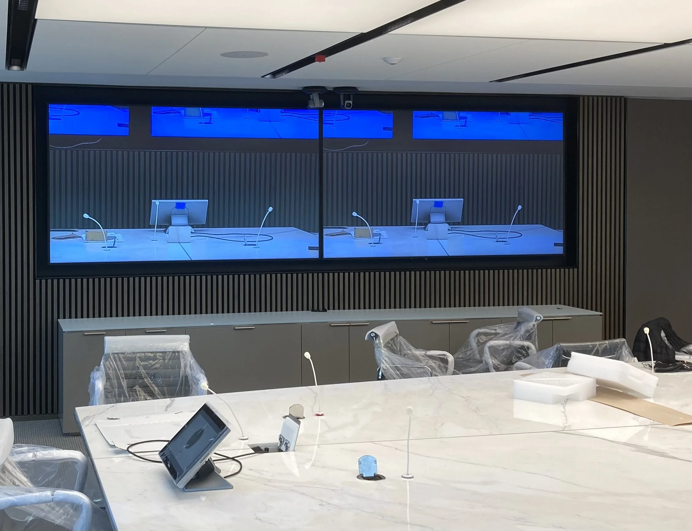 Modern conference room with a large white marble table, computers, and microphones, with a wall-mounted screen showing a digital blue background.