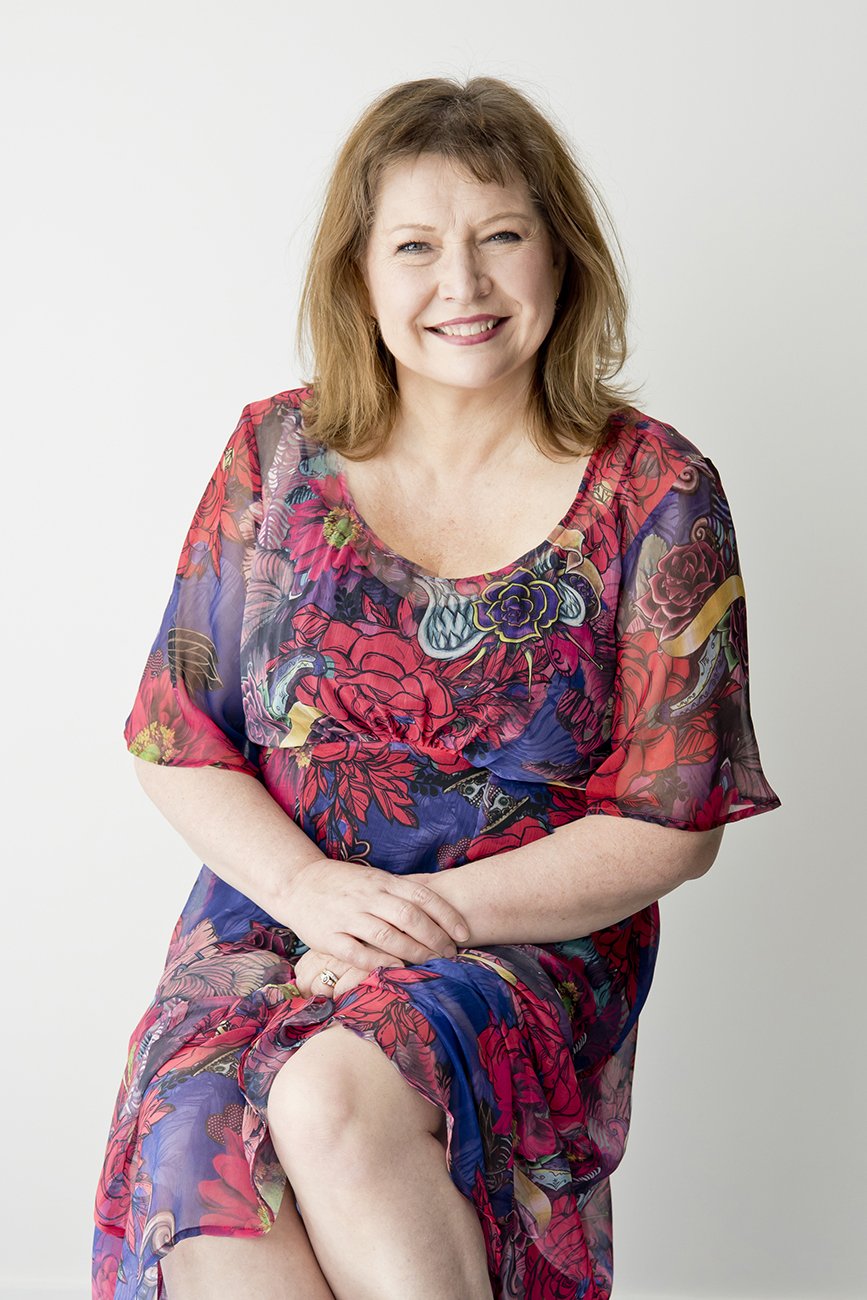 A woman with shoulder-length light brown hair, smiling, wearing a colorful floral dress, sitting with her legs crossed against a plain light gray background.