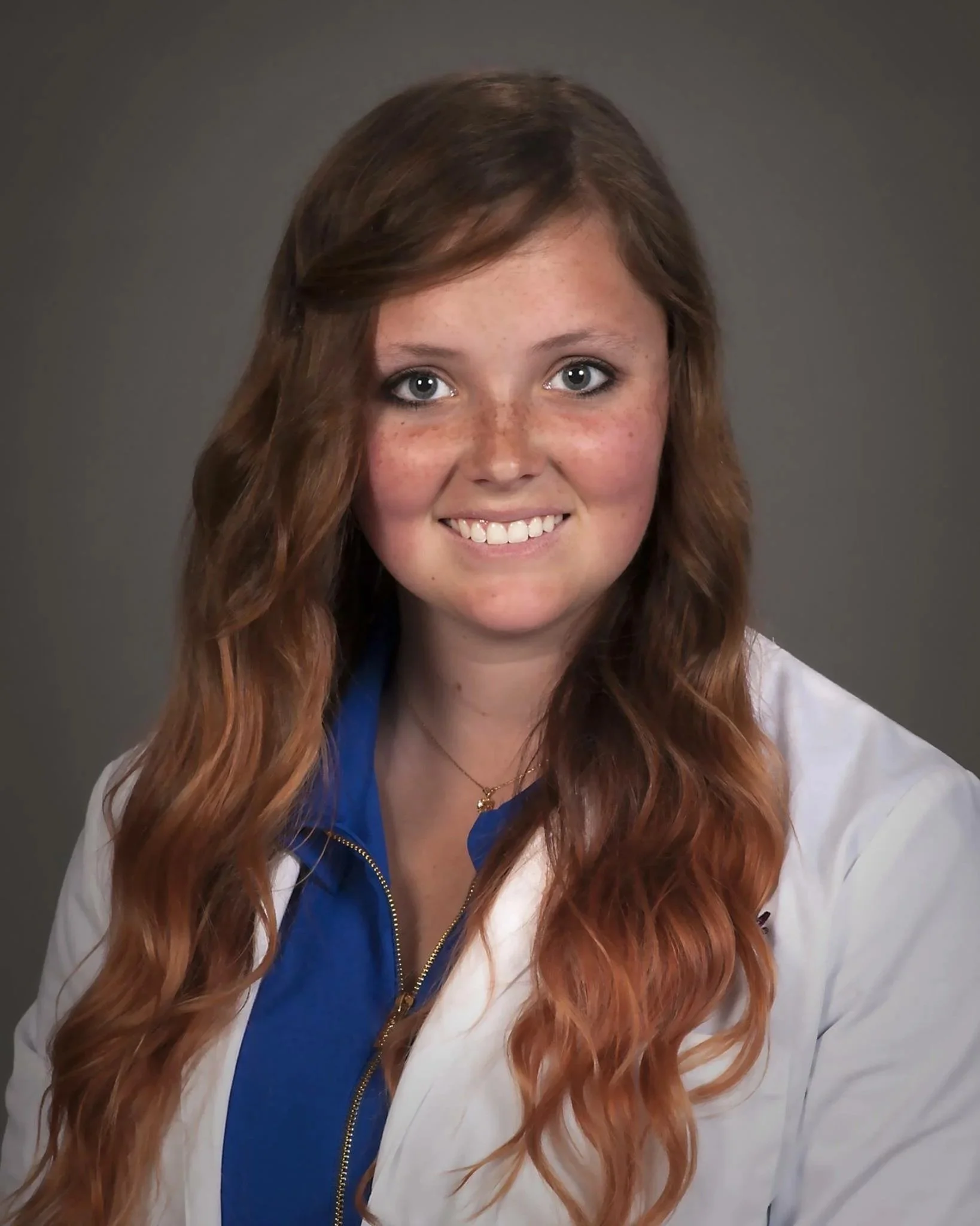 Close-up of a young woman with long red hair, gray eyes, freckles, smiling, wearing a blue shirt and white jacket, against a gray background.