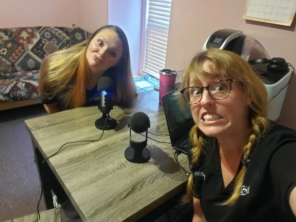 Two women making silly faces at a camera set up on a table with microphones, a laptop, and a dark pink travel mug in a room with pink walls and a window with blinds.