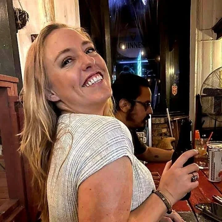 A smiling woman with long blonde hair in a white shirt, holding a drink in a bar or restaurant with other patrons, including a man with glasses in the background.