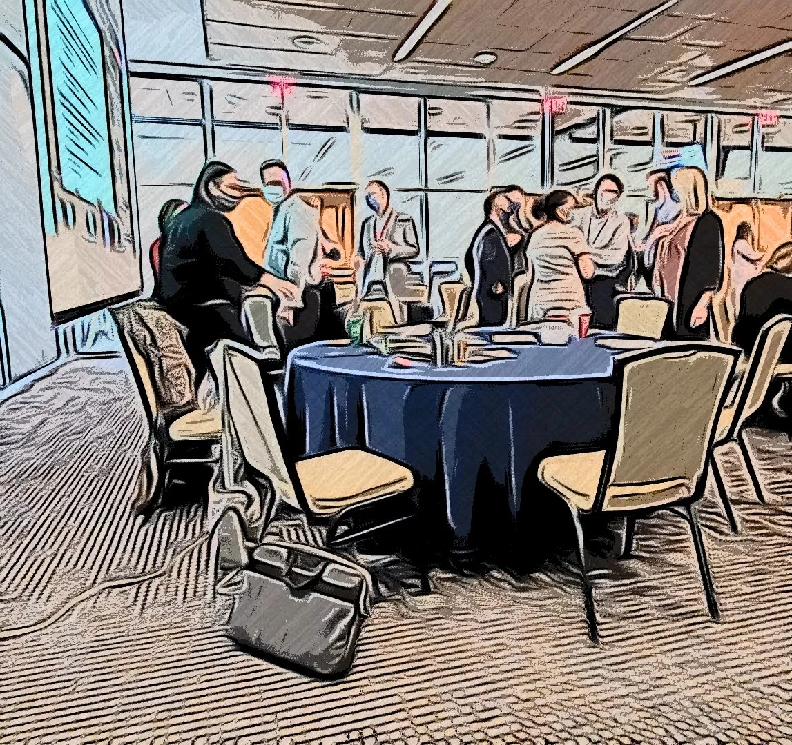 Group of people gathered in a conference room with large windows, standing and sitting around a table with glasses and bottles, engaged in conversation.