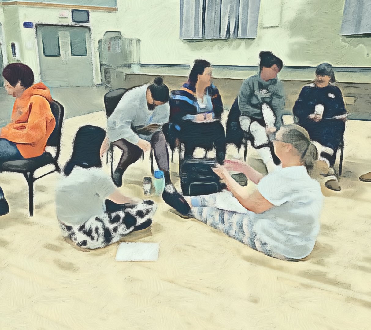 Group of six people sitting in chairs and on the floor, engaged in conversation in a room with a wooden floor and door in the background.
