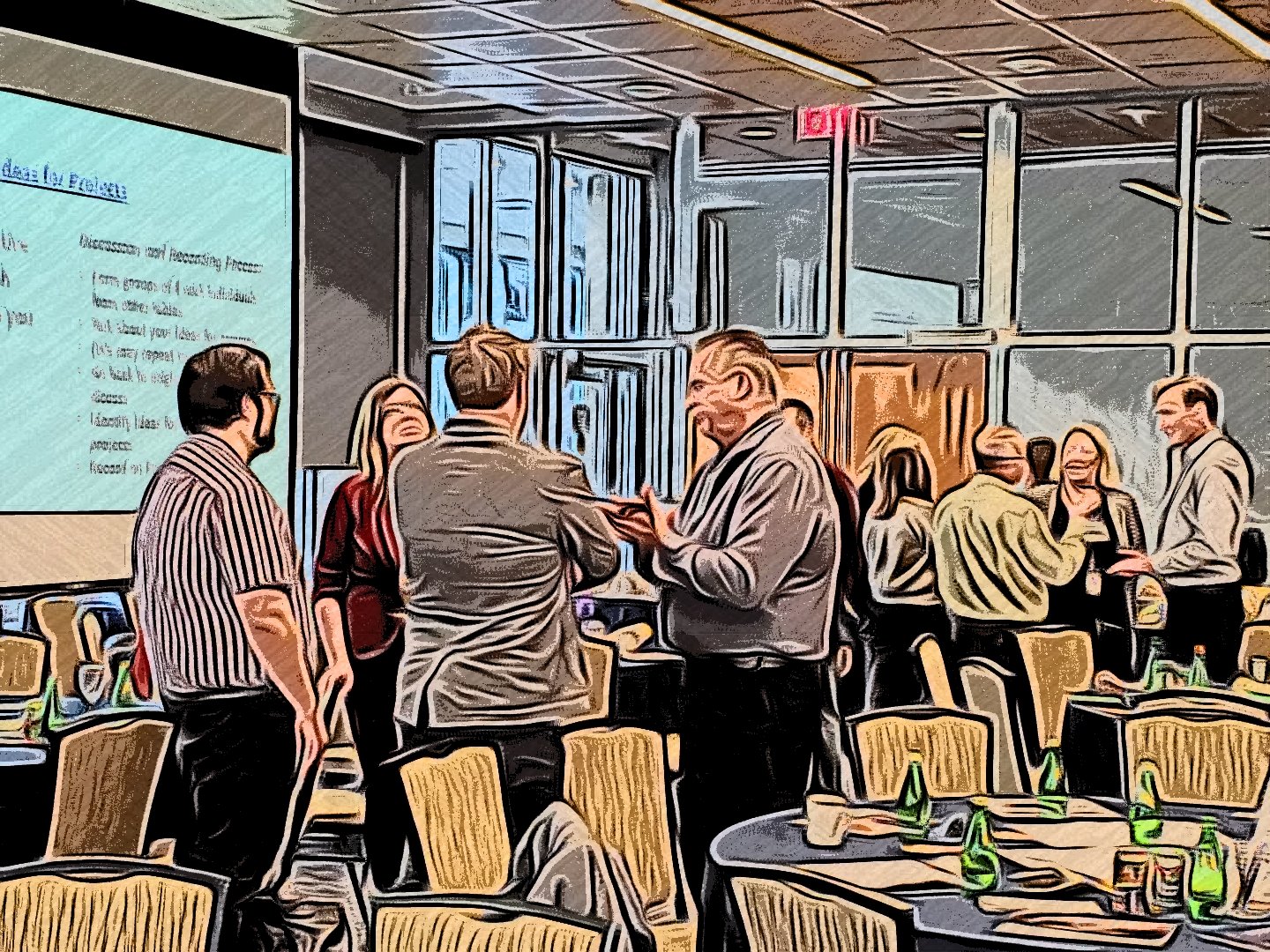 A group of people in business attire engaged in conversations in a conference room with tables, chairs, and a large screen displaying presentation slides.