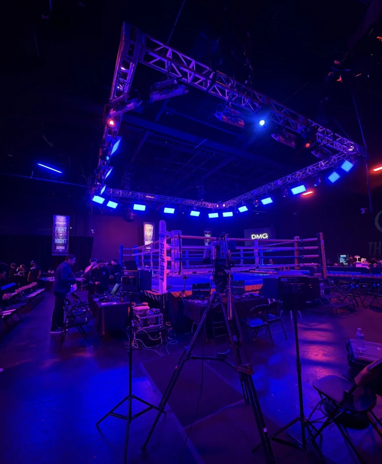 Photo of an empty boxing ring in a dark venue with blue and purple lighting, surrounded by seating and event equipment.