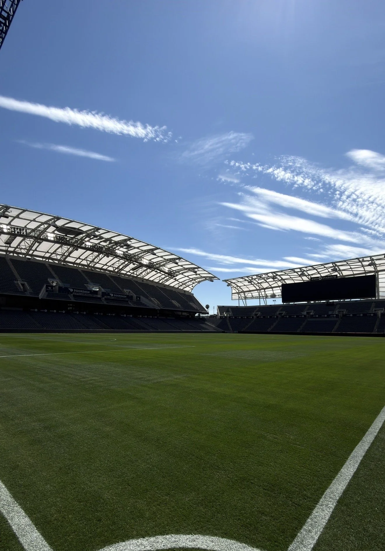 An empty soccer stadium with green grass field and modern roof structures, under a bright blue sky with scattered white clouds.