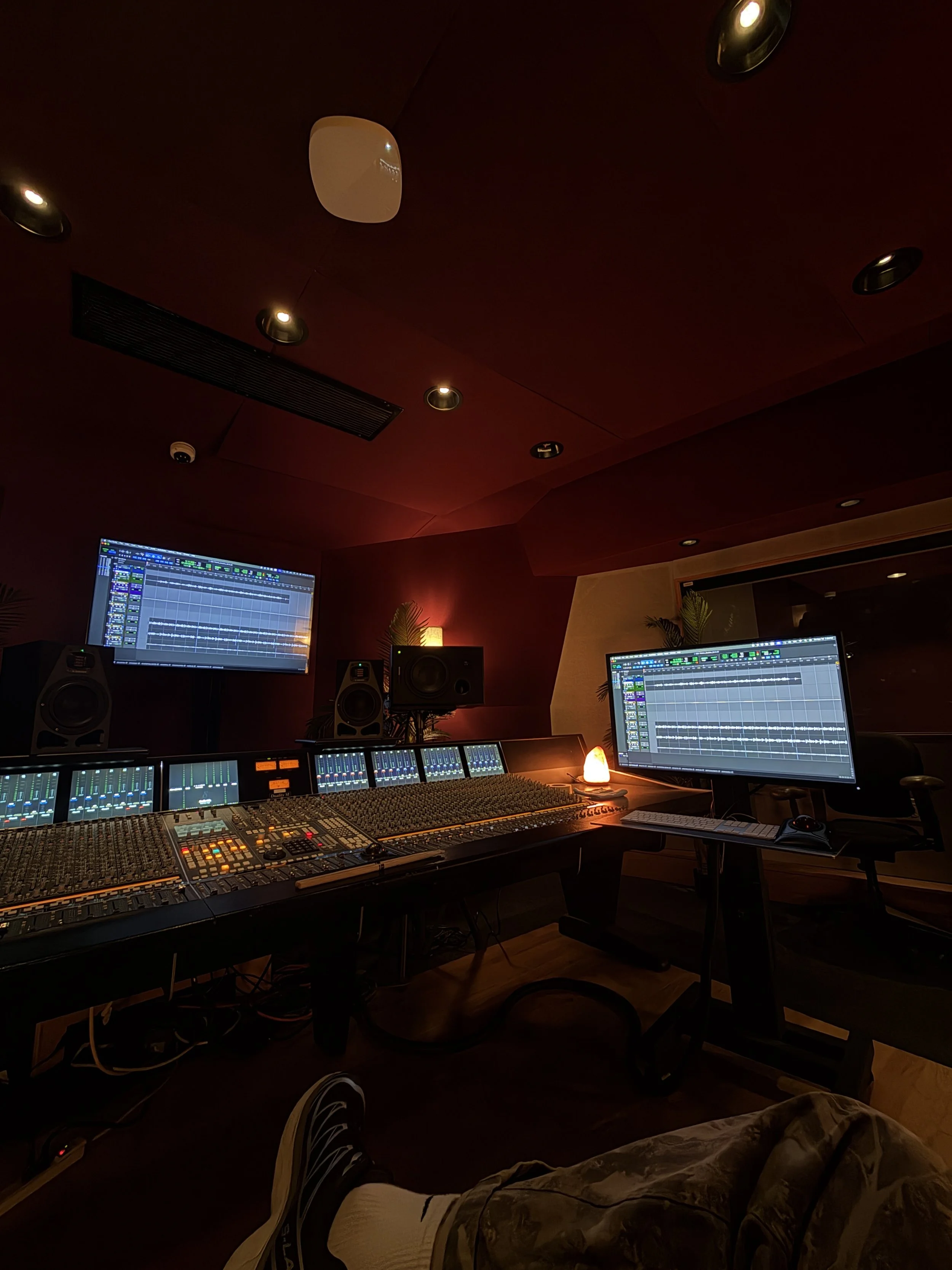 A music recording studio with a mixing console, multiple monitors displaying digital audio workstations, studio speakers, and framed by a person in camouflage pants and sneakers relaxing nearby.