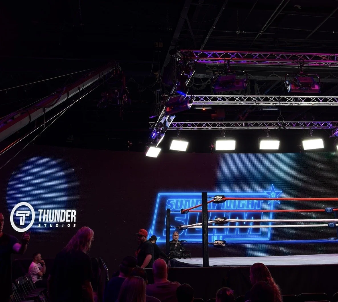 Inside a dark wrestling arena with a wrestling ring illuminated, featuring Bright neon signs that say "Saturday Night Slam" and a logo for Thunder Studios, with audience members waitng around.