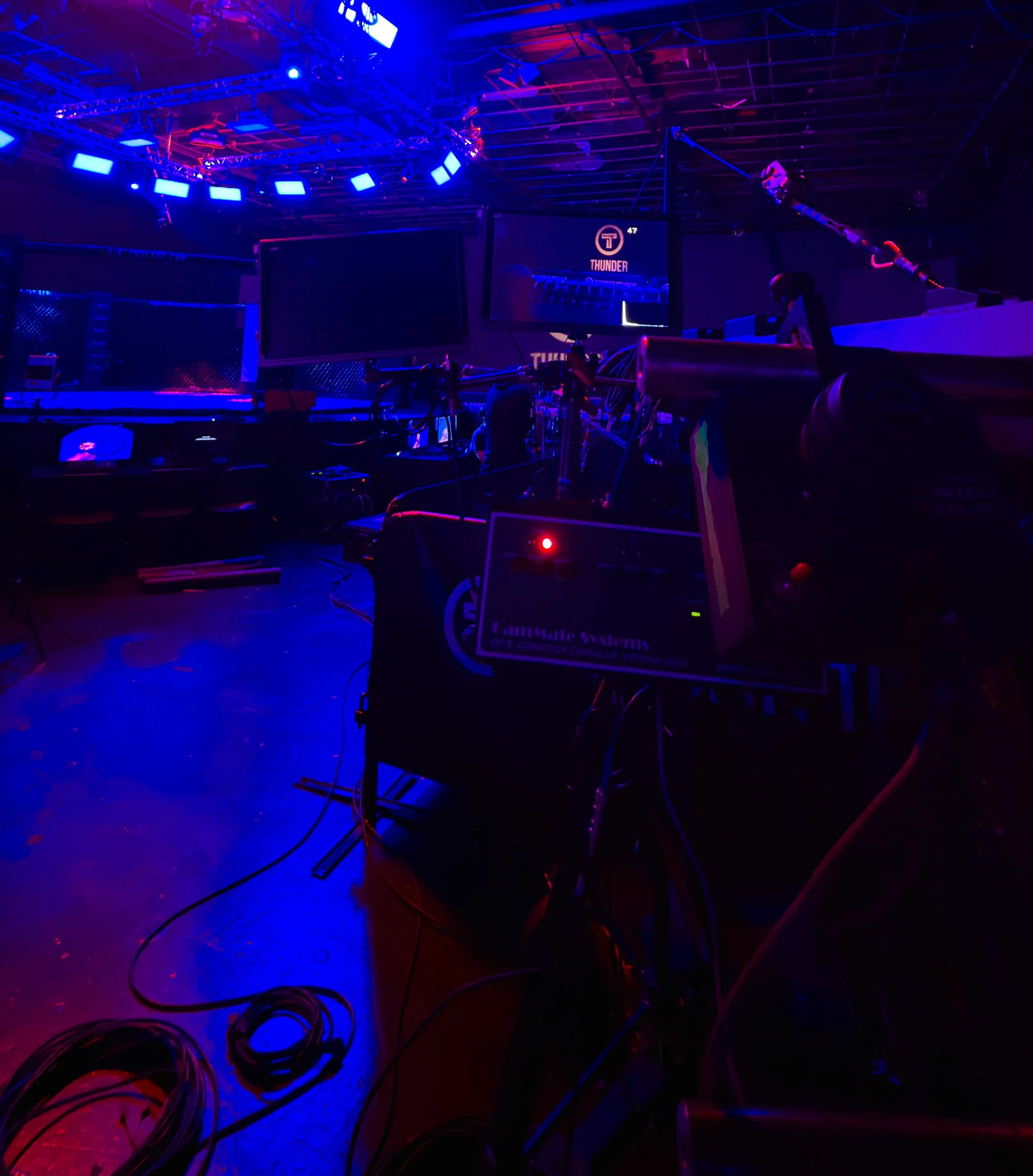 Photo of a dark room illuminated by blue and purple LED lights, with computer monitors, wires, and electronic equipment, suggesting a setup for a live performance or gaming event.