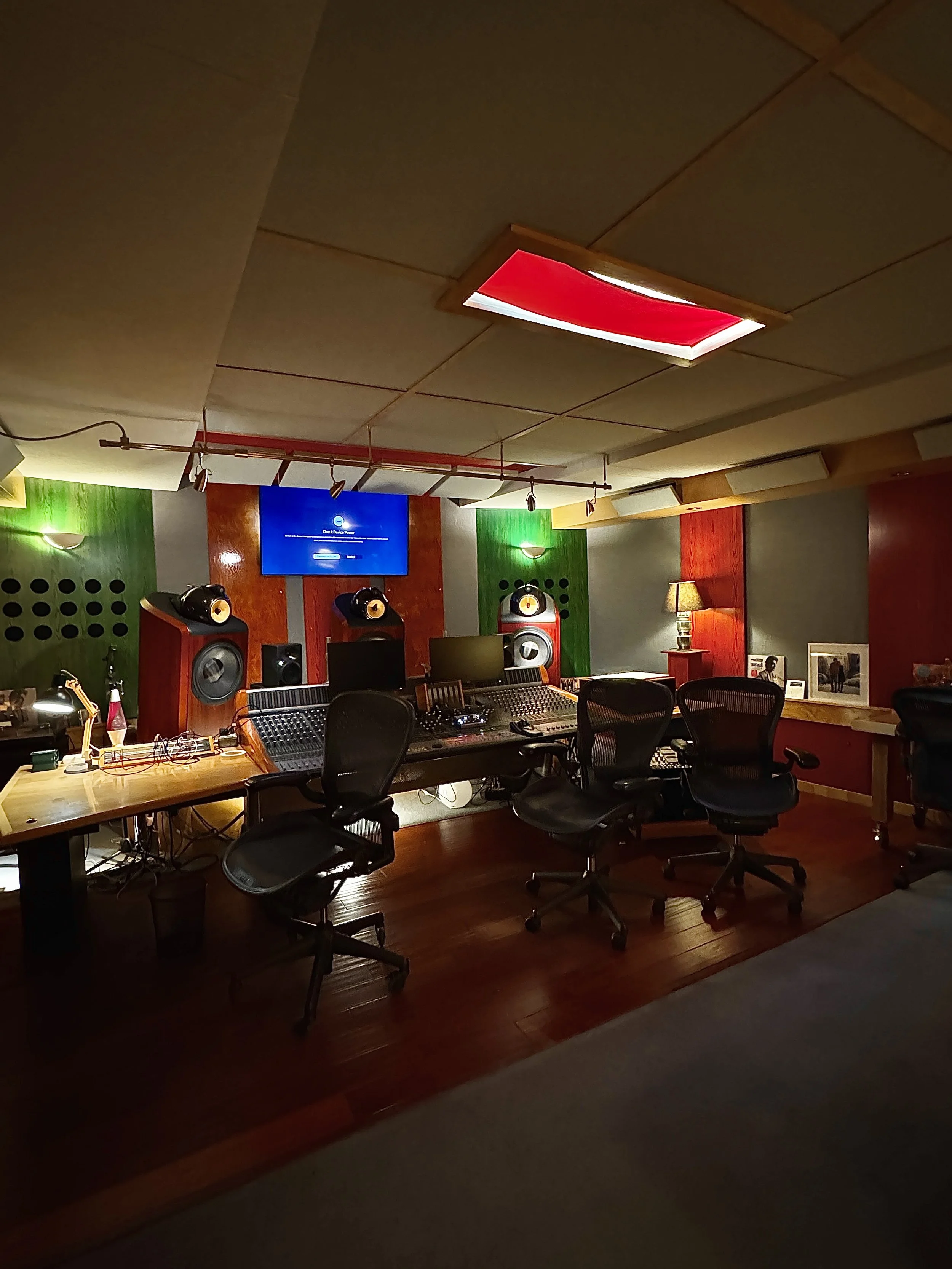 Music recording studio with mixing console, studio monitors, and a large screen, featuring wooden flooring and acoustic panels on the walls.