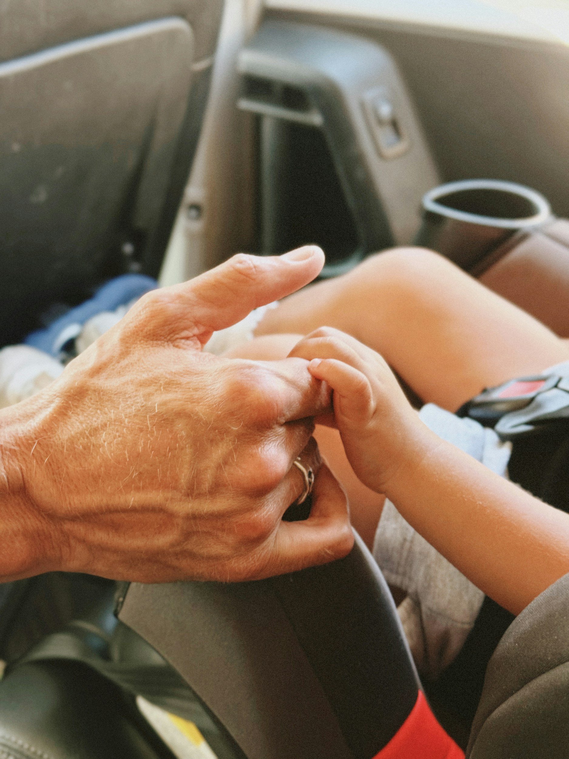A close-up of an adult's hand gently holding a child's hand, seated next to each other in a vehicle. The adult's hand has a ring and the child's hand is smaller. The seating area and cup holder of the vehicle are visible in the background.