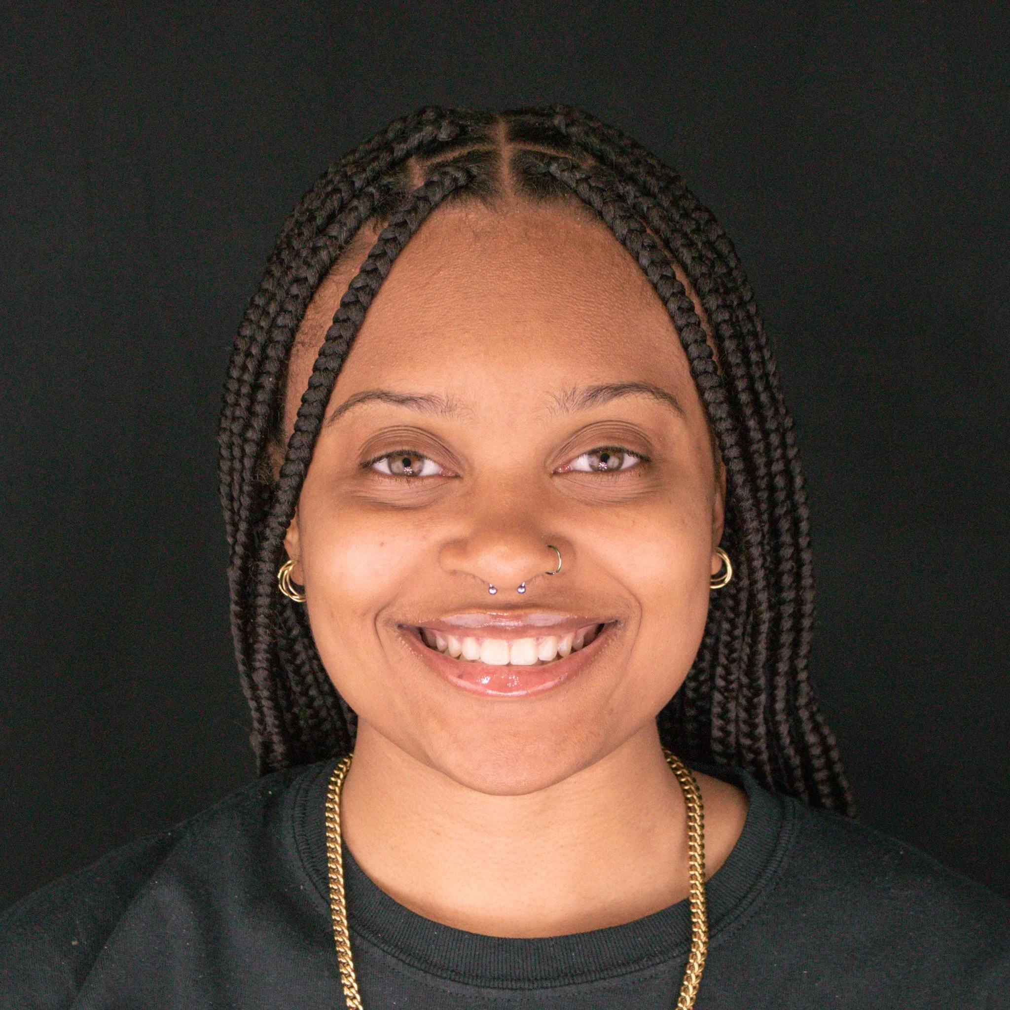 Close-up of a smiling woman with braided hair, wearing gold jewelry, against a black background.