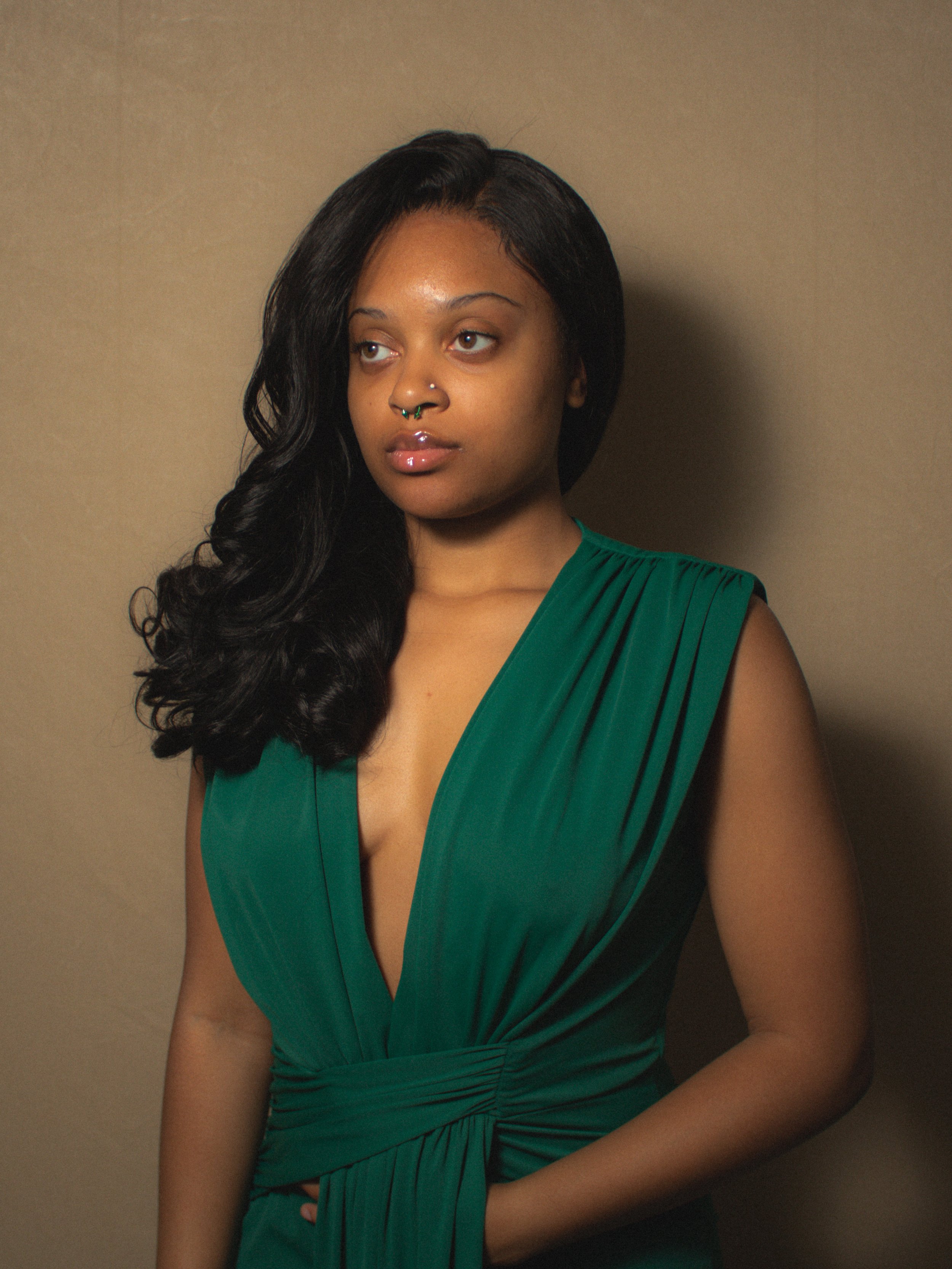 A woman with long, wavy black hair in a side part, wearing a green sleeveless dress with pleats, standing against a plain background.